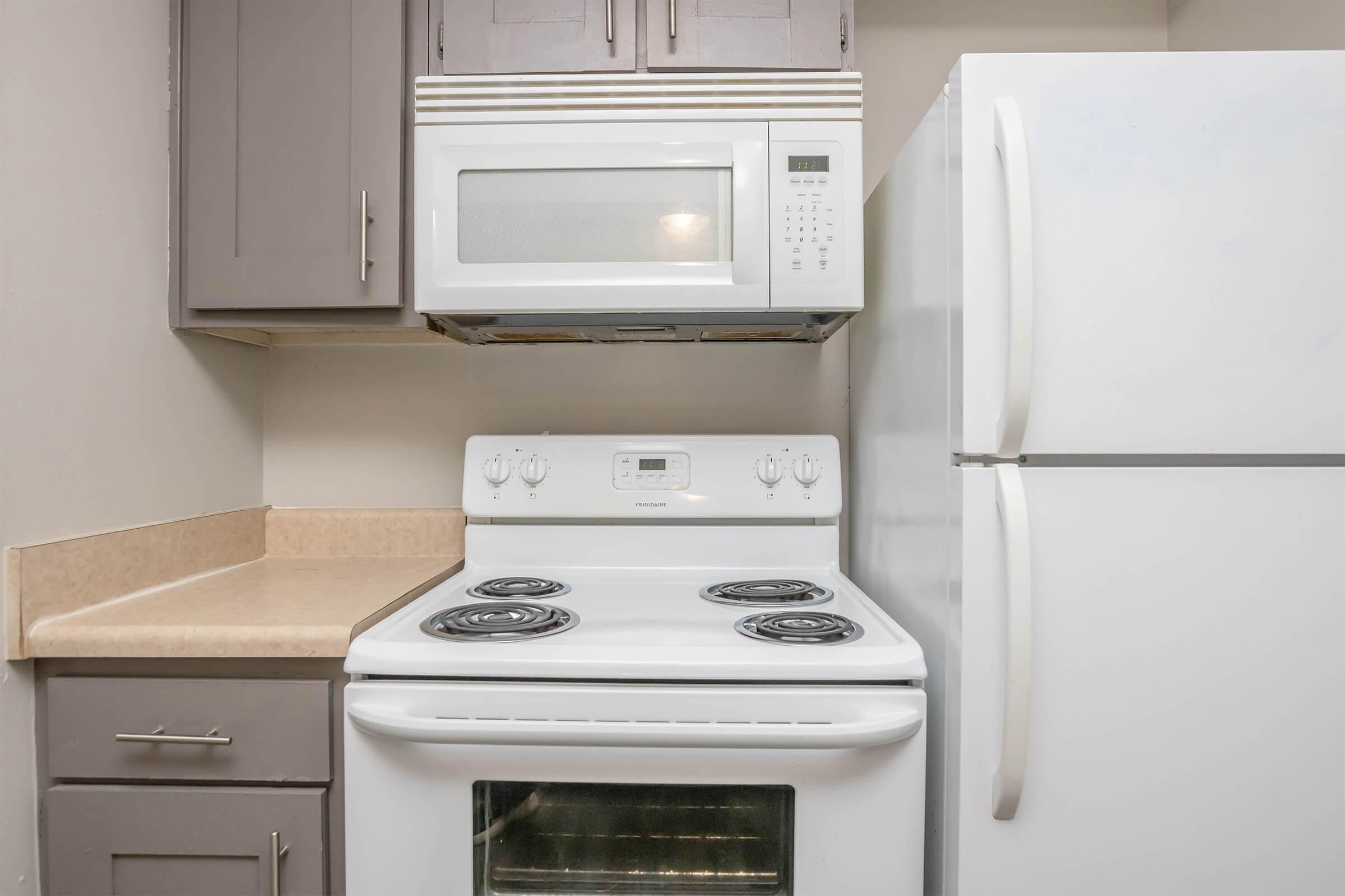 A compact kitchen featuring a white stove with four burners, a microwave situated above the stove, and a white refrigerator. The cabinetry is gray, and there is a light-colored countertop. The overall layout is functional and efficient for small space cooking.