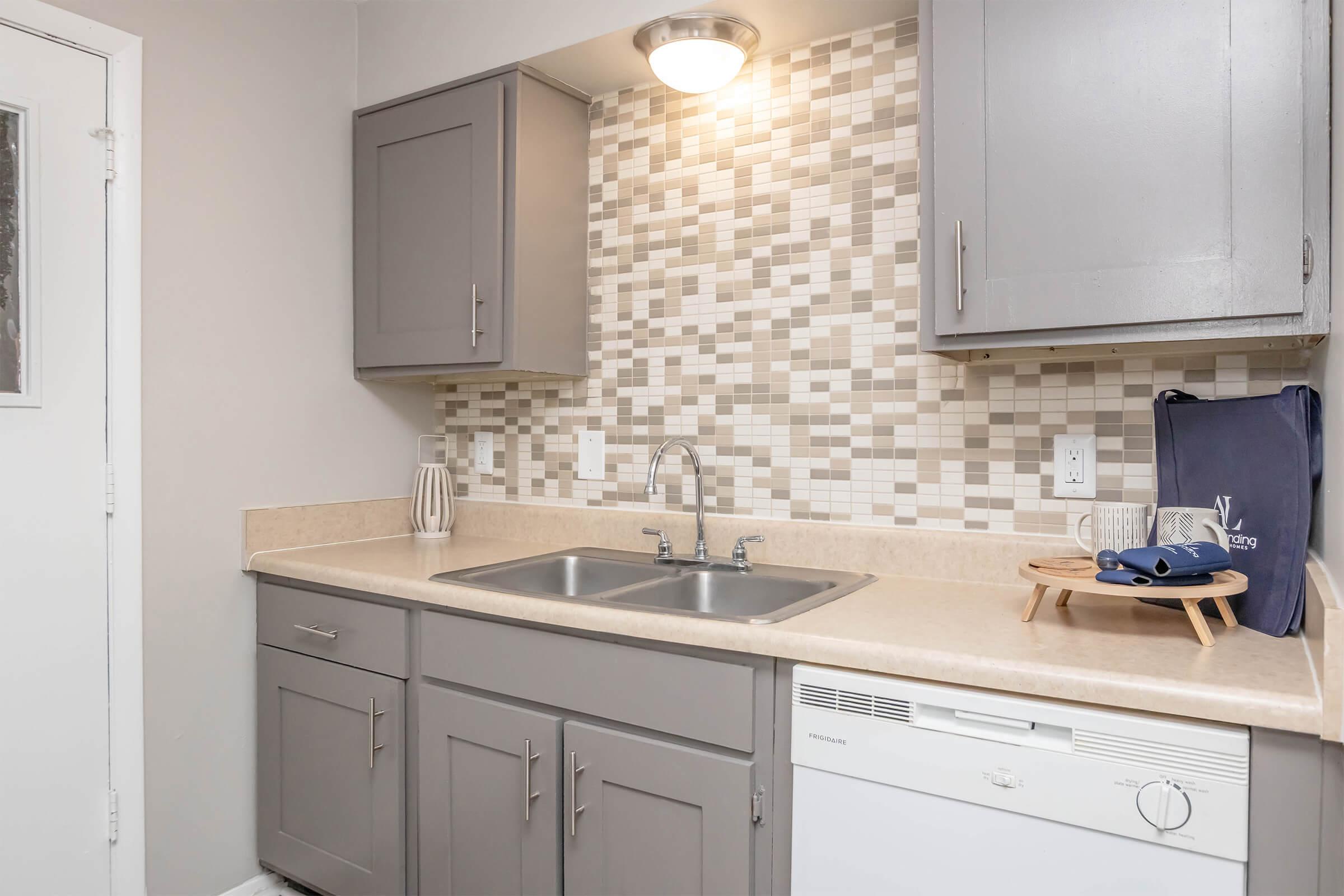 Modern kitchen featuring grey cabinets, a double sink, and a beige countertop. The backsplash consists of small, multicolored tiles. To the right, there’s a dishwasher, and on the countertop, there are glasses and a serving tray. A door is visible on the left.