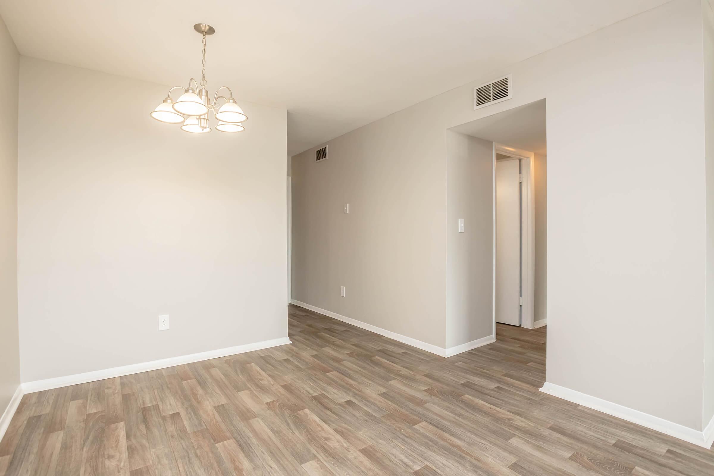 A well-lit, empty living space featuring a light fixture with multiple bulbs, light gray walls, and a wood-style floor. To the right, there is an open doorway leading to a hallway. The overall atmosphere appears spacious and inviting.