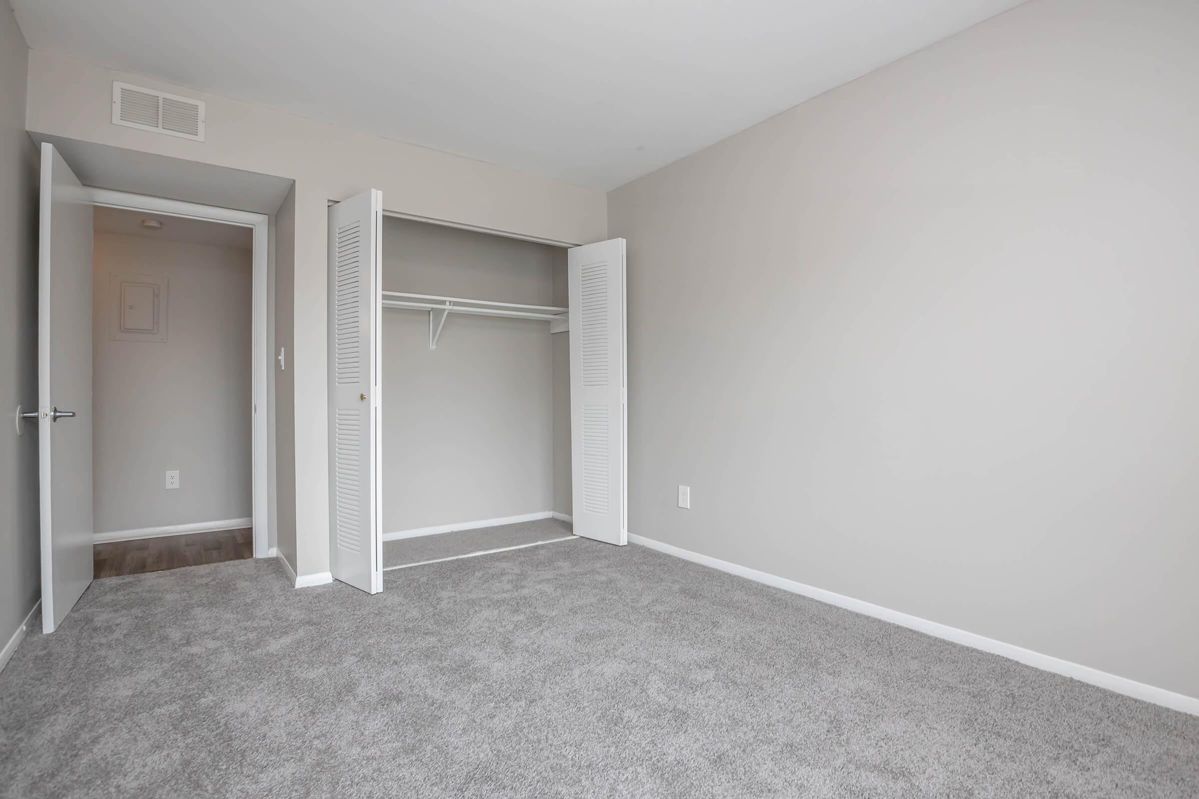 A vacant room with light gray walls and carpeted flooring. There is a closet with open double doors on the left and a doorway leading to another space on the left. The room has ample natural light and a clean, minimalist appearance.