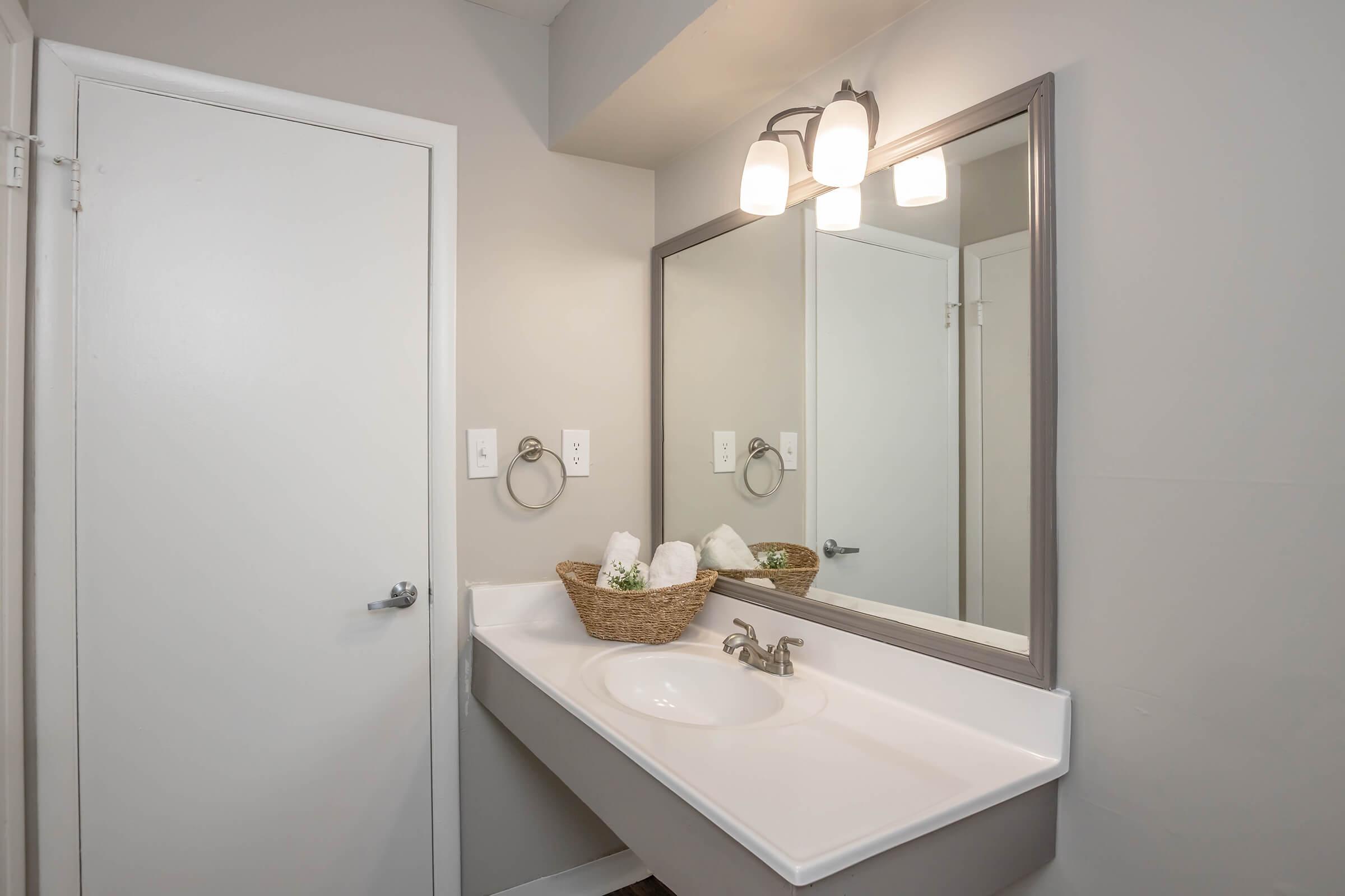 A modern bathroom featuring a white sink with a silver faucet, a large mirror above it, and a light fixture with three bulbs. There's a wooden basket on the counter holding rolled towels. The walls are painted a light gray, and there's a white door and towel ring on the right.