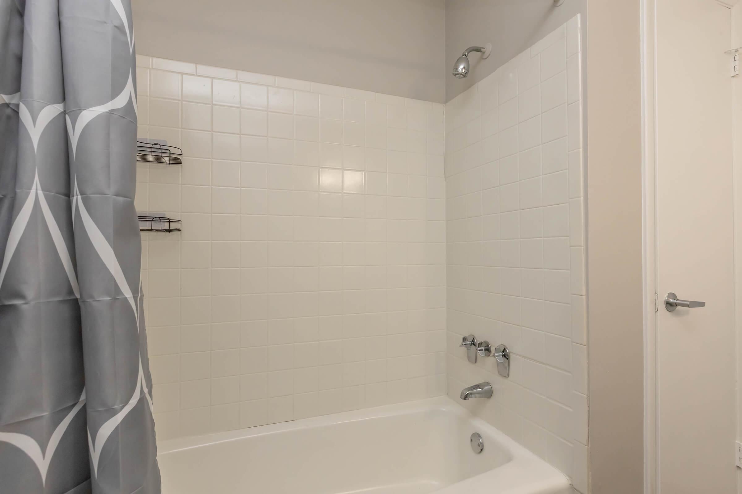 A clean and modern bathroom featuring a white tiled shower and tub area, with a gray patterned shower curtain and chrome fixtures. Two wire shelves are mounted on the wall, providing storage space. The overall aesthetic is bright and minimalist, with neutral tones.