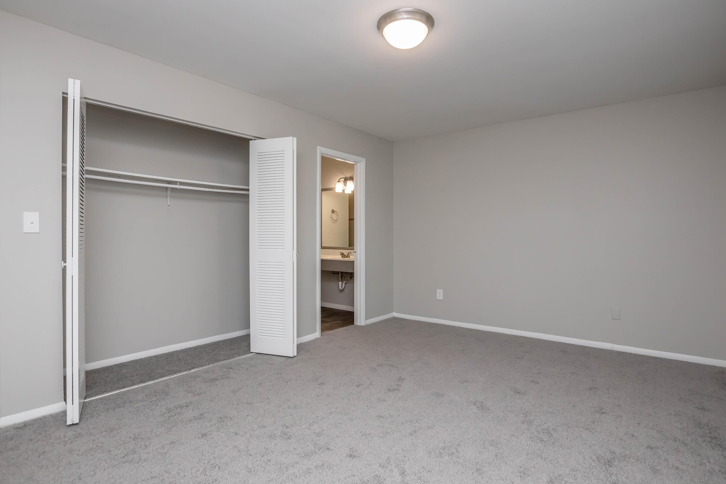 A spacious, empty bedroom featuring light gray walls and carpeting. There is a closed closet with white louvered doors on one side. A doorway leads to a bathroom area with soft lighting. The room is well-lit from a central ceiling light, emphasizing a clean and minimalist space.