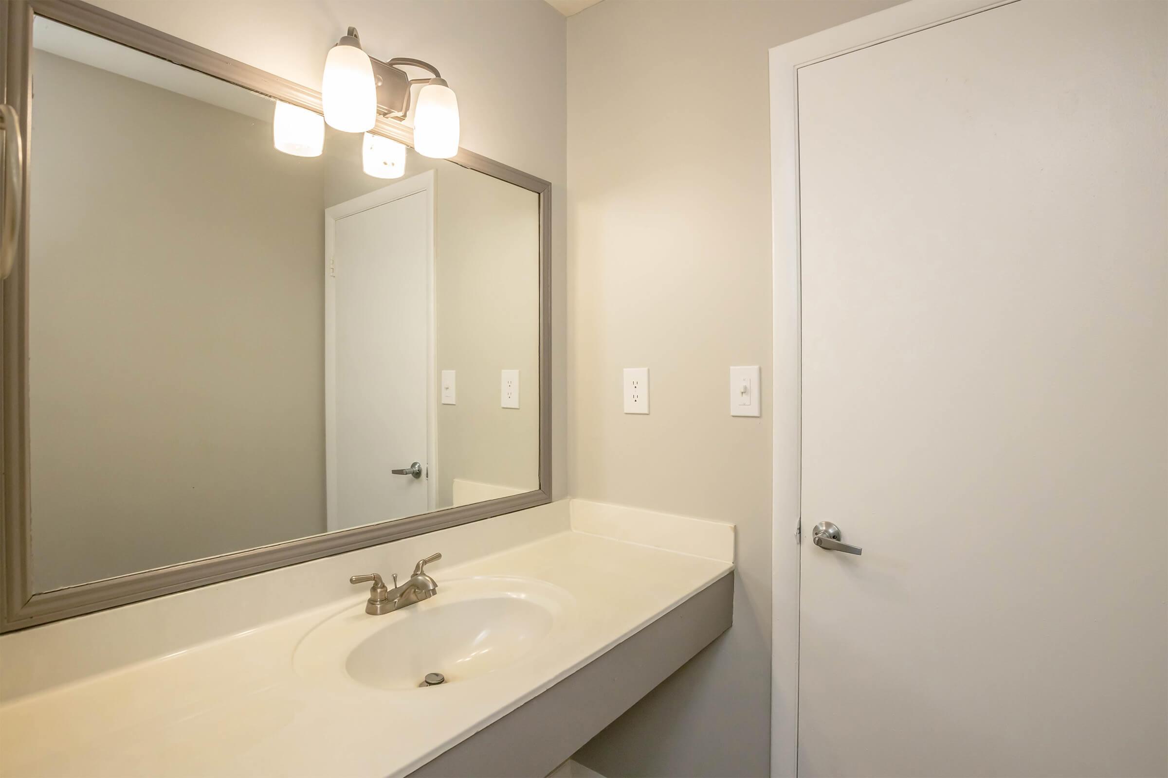A clean and modern bathroom with a light-colored countertop, a single sink, and a large mirror above it. The walls are painted in a neutral tone, and there are two light fixtures above the mirror. A closed white door is visible on the right side, enhancing the simple and uncluttered design of the space.