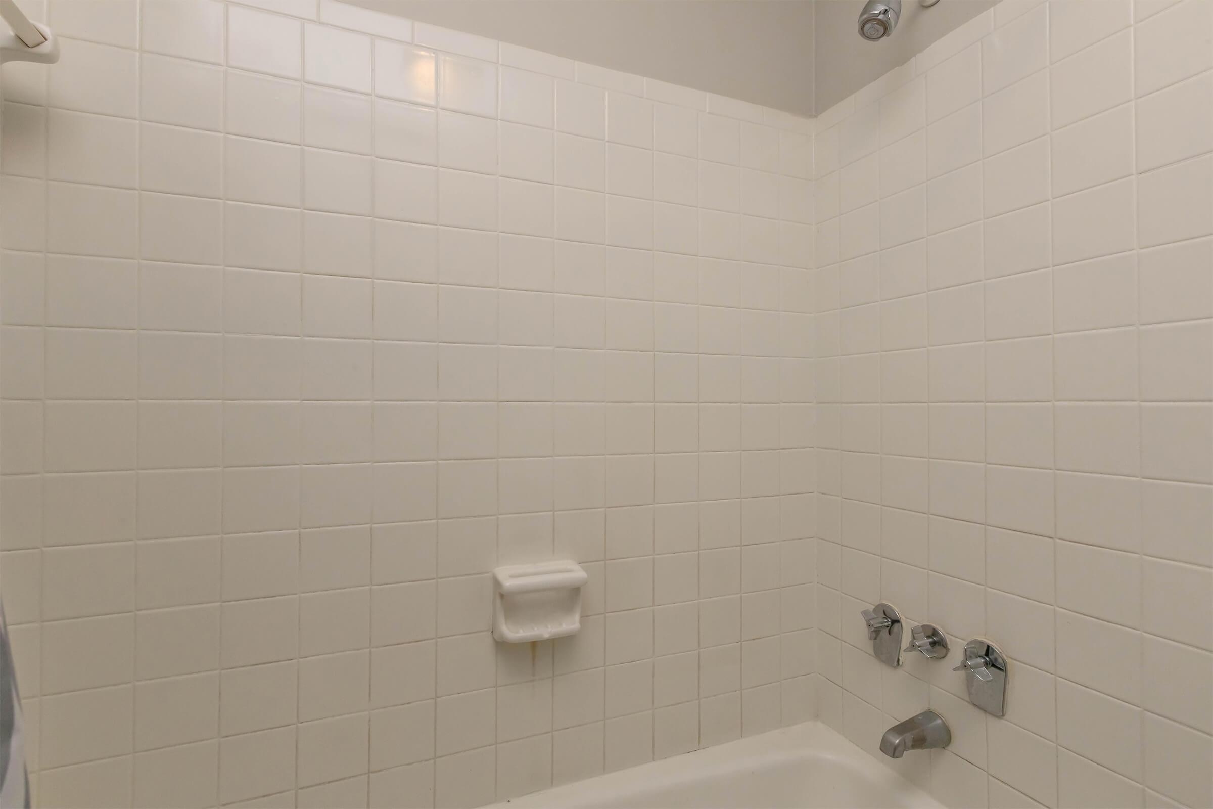 A clean and simple bathroom shower with white tiled walls and a silver faucet. There are two knobs for temperature control and a soap dish on the wall. The space appears well-lit and organized, with a minimalistic design.