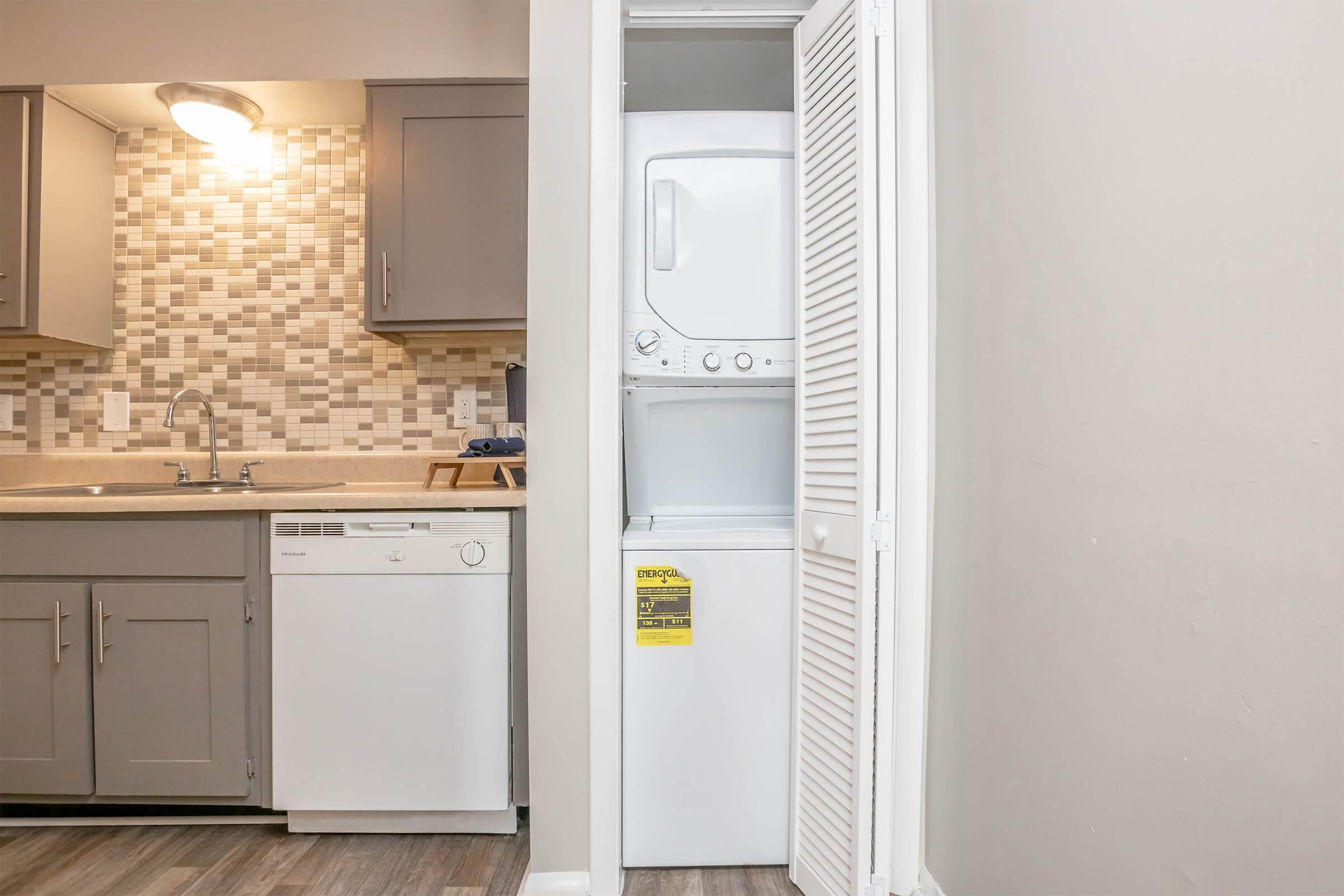 A compact kitchen featuring gray cabinets and a mosaic backsplash. A stacked washer and dryer is located in a closet next to the dishwasher, with a light illuminating the workspace. The flooring is a light wood laminate, enhancing the modern aesthetic of the kitchen.