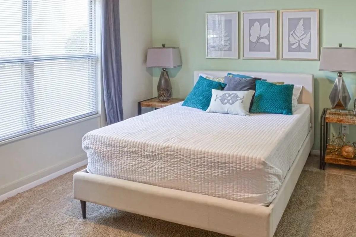 A neatly arranged bedroom featuring a light-colored bed with decorative pillows, two nightstands with lamps on either side, and three framed botanical prints on the wall. The room has pale green walls and natural light coming through a window, creating a calm and inviting atmosphere.