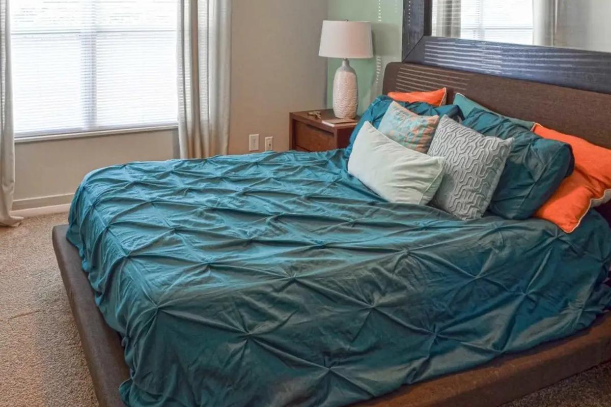 A neatly made bed with a teal quilt and several colorful pillows in a cozy bedroom. There is a bedside table with a lamp and a window with sheer curtains, allowing natural light to brighten the space. The carpeted floor adds to the warm, inviting atmosphere of the room.
