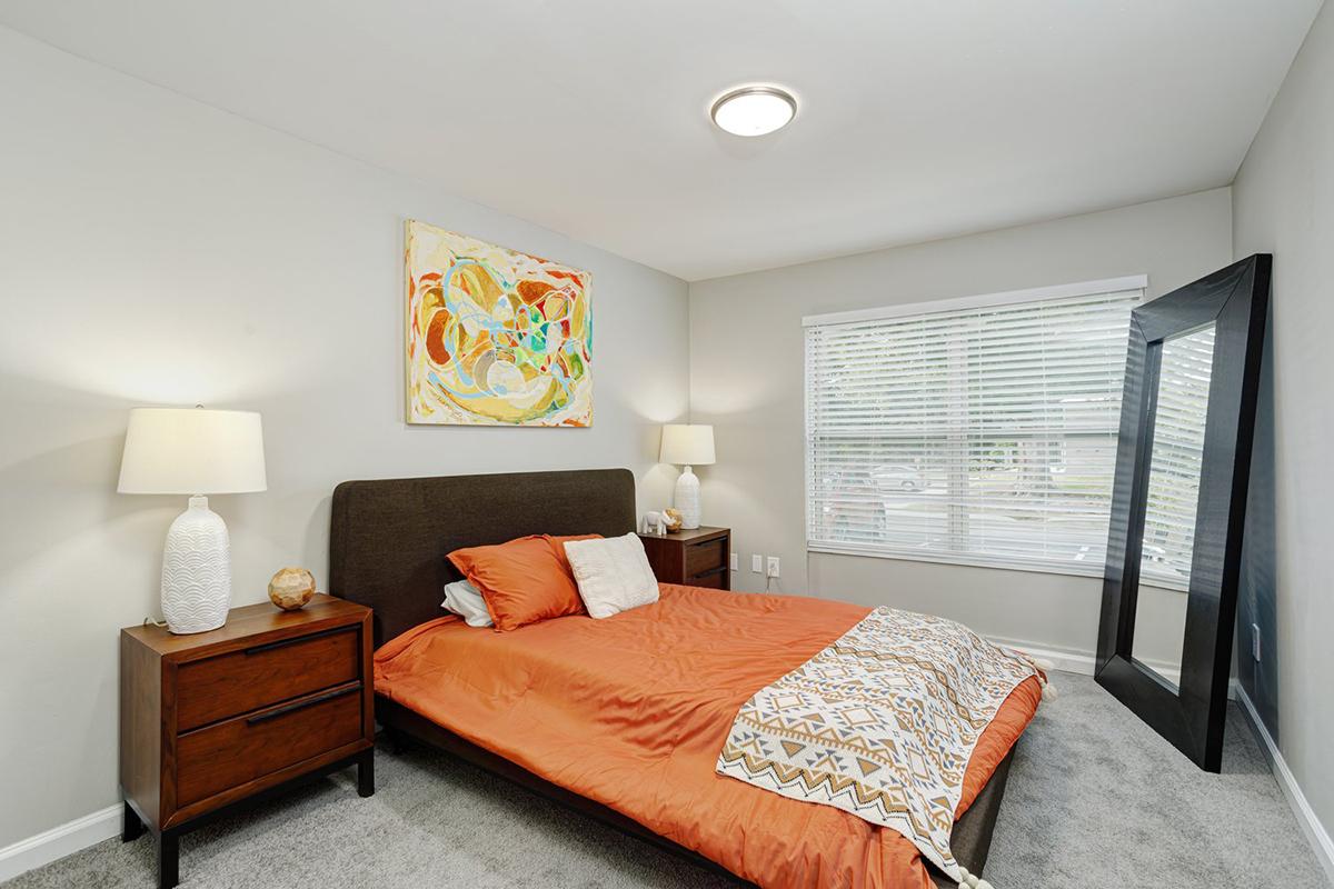 A modern bedroom with an orange bedspread and coordinating pillows on a queen-sized bed. There are two bedside tables with lamps, a large mirror against the wall, and a colorful abstract painting above the bed. Natural light floods in through a window with white blinds, illuminating the neutral-toned walls and carpet.