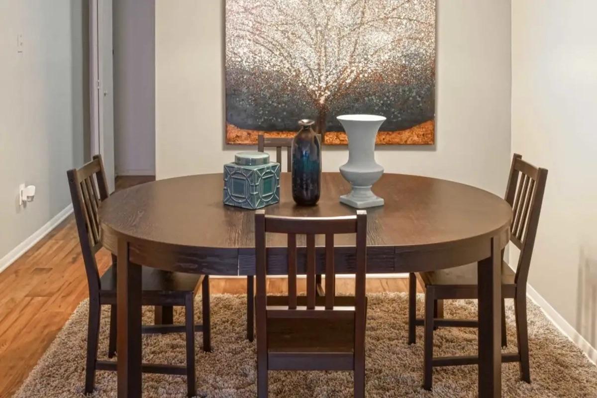 A round wooden dining table with four chairs is positioned on a plush rug. Centered on the table are decorative items: a geometric teal box, a tall dark blue vase, and a white sculptural vase. Behind the table, a large artwork depicting a tree fills the wall. The setting has a warm, inviting atmosphere.