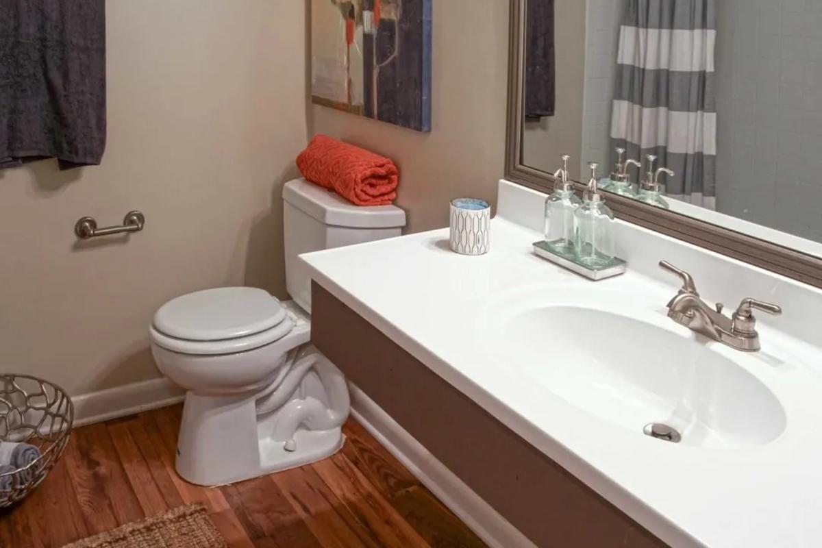 A clean and modern bathroom featuring a white sink and toilet, a large mirror above the sink, and decorative items on the countertop. The walls are painted a neutral color, and a striped shower curtain is visible. A basket and rolled towels add to the decor. Wooden flooring completes the warm ambiance.