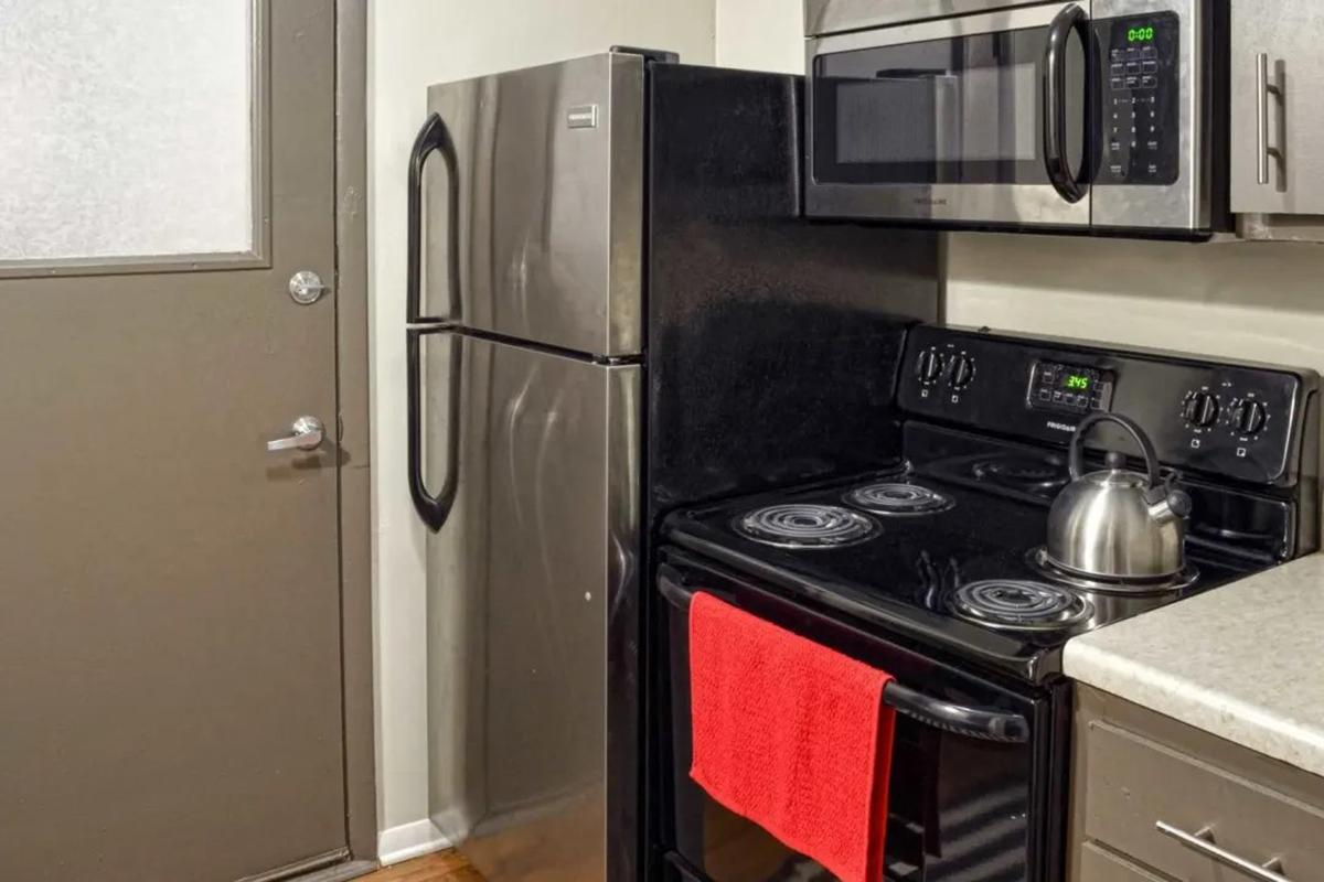 A modern kitchen featuring a black refrigerator and stove. The stove has a kettle on it and a red kitchen towel hanging from the oven handle. There is a door in the background, with a frosted glass panel, and the kitchen has neutral-colored cabinets and a light countertop.