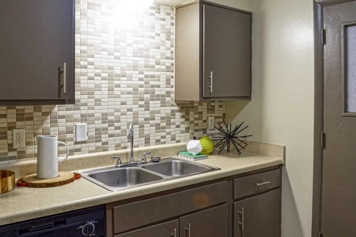 A modern kitchen featuring a beige countertop, double sink, and dark cabinets. The backsplash is decorated with mosaic tiles in various shades. A white pitcher, a green decorative item, and a star-shaped centerpiece are placed on the counter. A door is visible on the right side of the image.