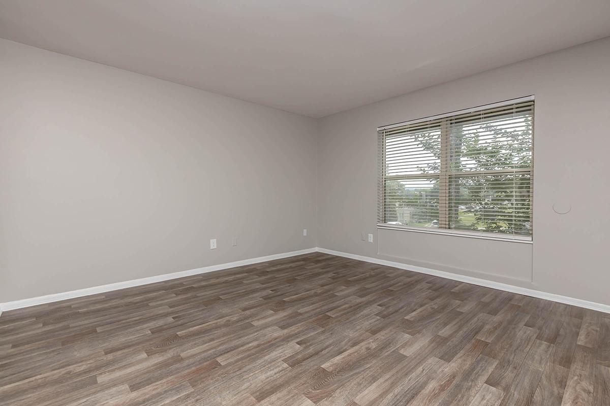 A spacious, empty room with light gray walls and a large window featuring horizontal blinds. The floor is covered with brown laminate wood, and there's a view of greenery outside the window. The room is bright and airy, suitable for various purposes.