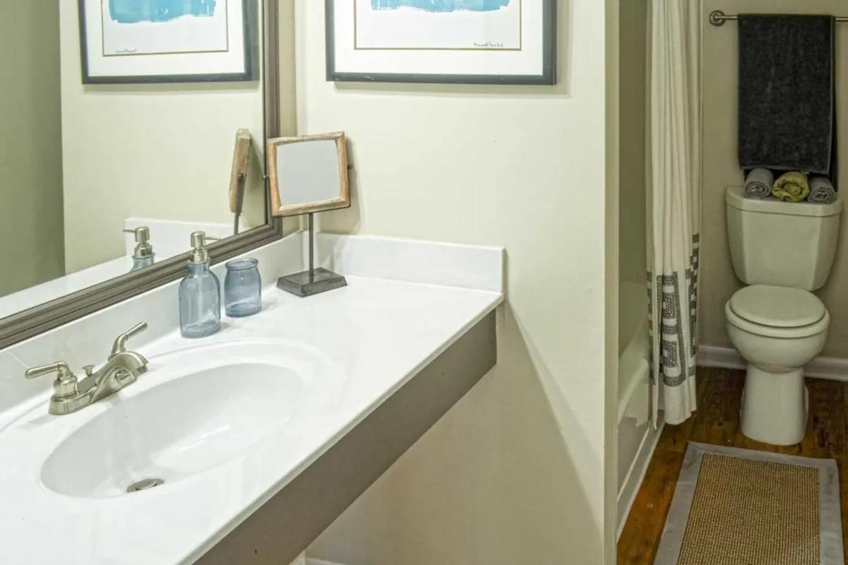 A clean, modern bathroom featuring a white sink with a faucet, two blue glass jars beside it, a rectangular mirror above the sink, and a shower curtain. To the right, a toilet is visible, accompanied by neatly arranged towels. The wooden floor adds a warm touch to the space.