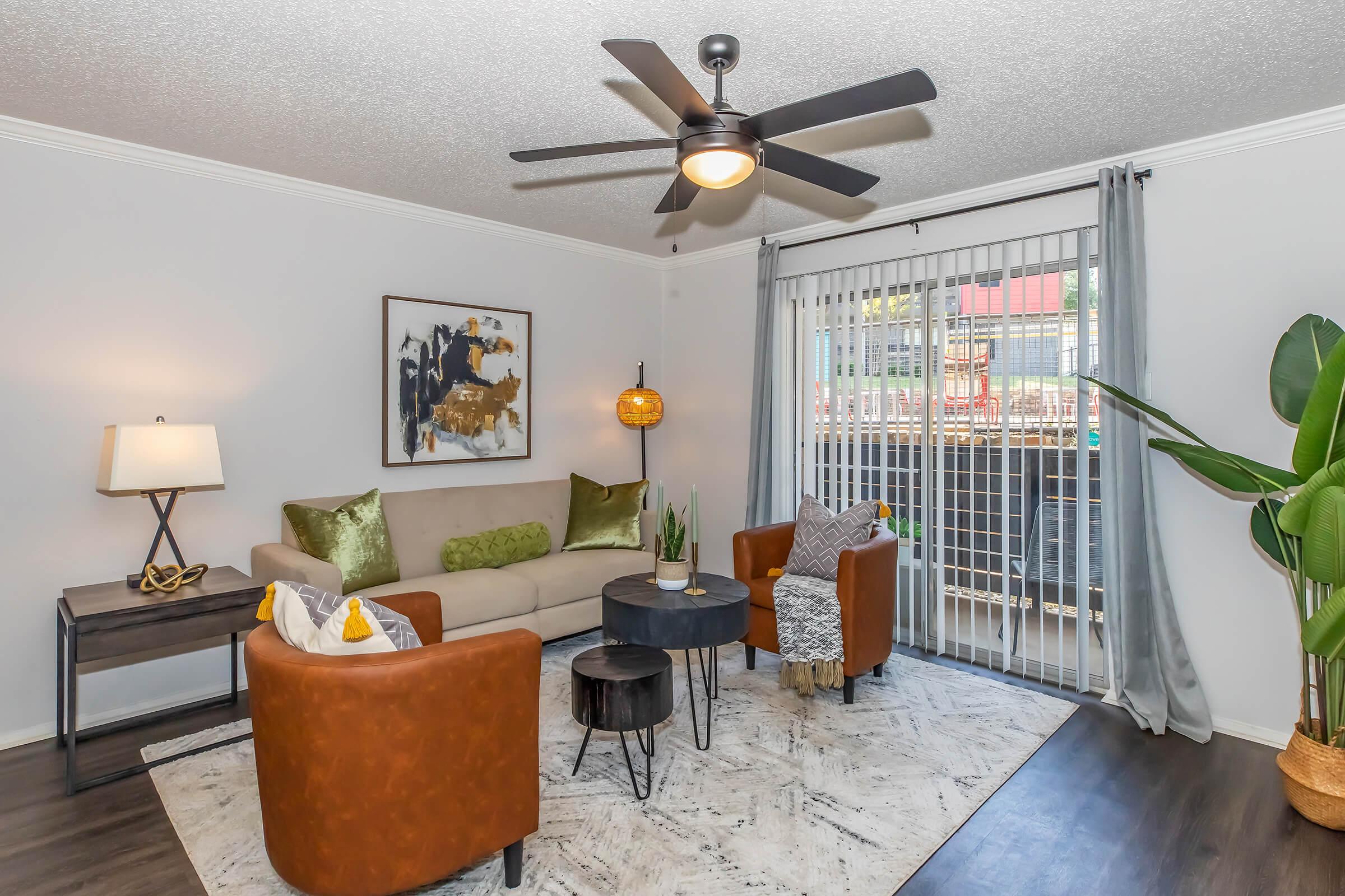 A cozy living room featuring a beige sofa with decorative pillows, a round black coffee table, and a pair of brown armchairs. A modern ceiling fan hangs above, and large sliding glass doors lead to an outdoor space. There is a plant in the corner and soft lighting from a lamp on a side table.