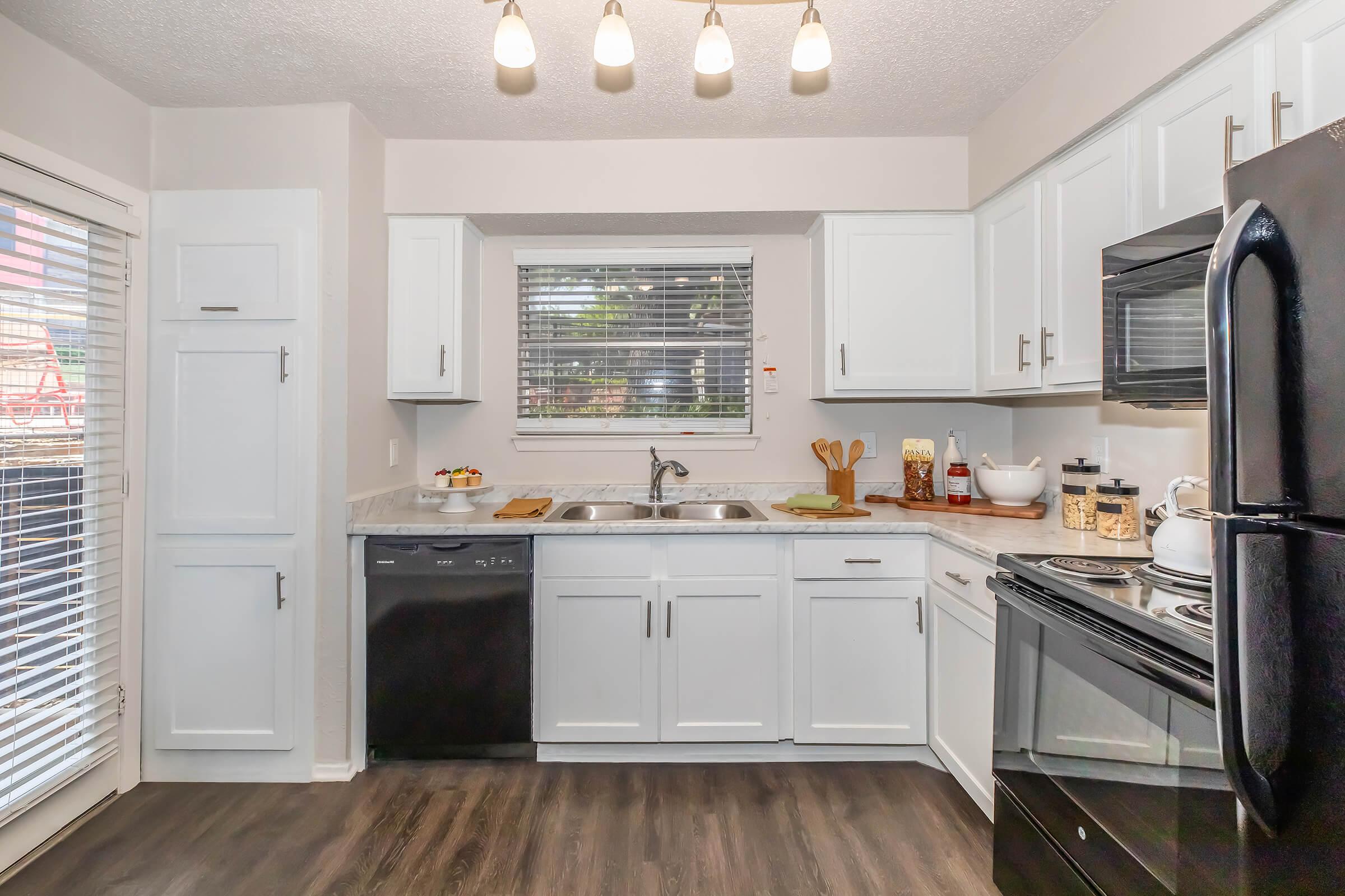 A modern kitchen featuring white cabinetry, a black refrigerator, a black stove, and a double sink under a window. Natural light filters through the window, and the countertops are decorated with small kitchen items, including a bowl of fruit. The flooring is dark wood, adding contrast to the light cabinets.