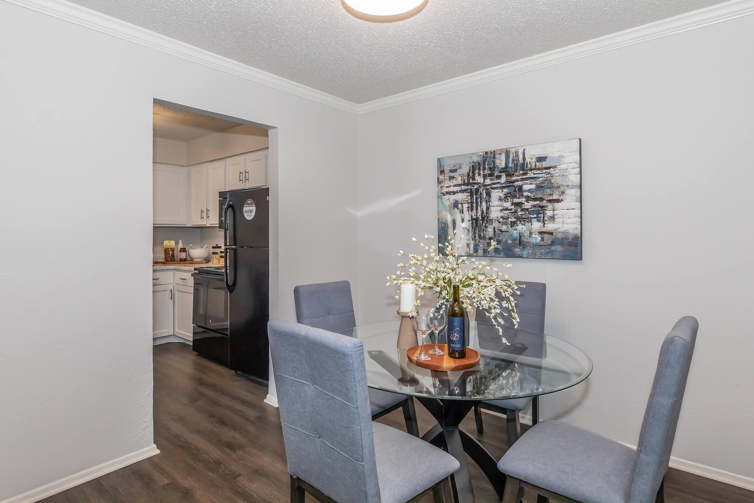 A modern dining area featuring a round glass table surrounded by four gray upholstered chairs. A vase with flowers sits on the table, and a candle is placed nearby. In the background, a kitchen is visible, showcasing white cabinets and a black refrigerator. The walls are painted in a light color, creating a bright, inviting atmosphere.