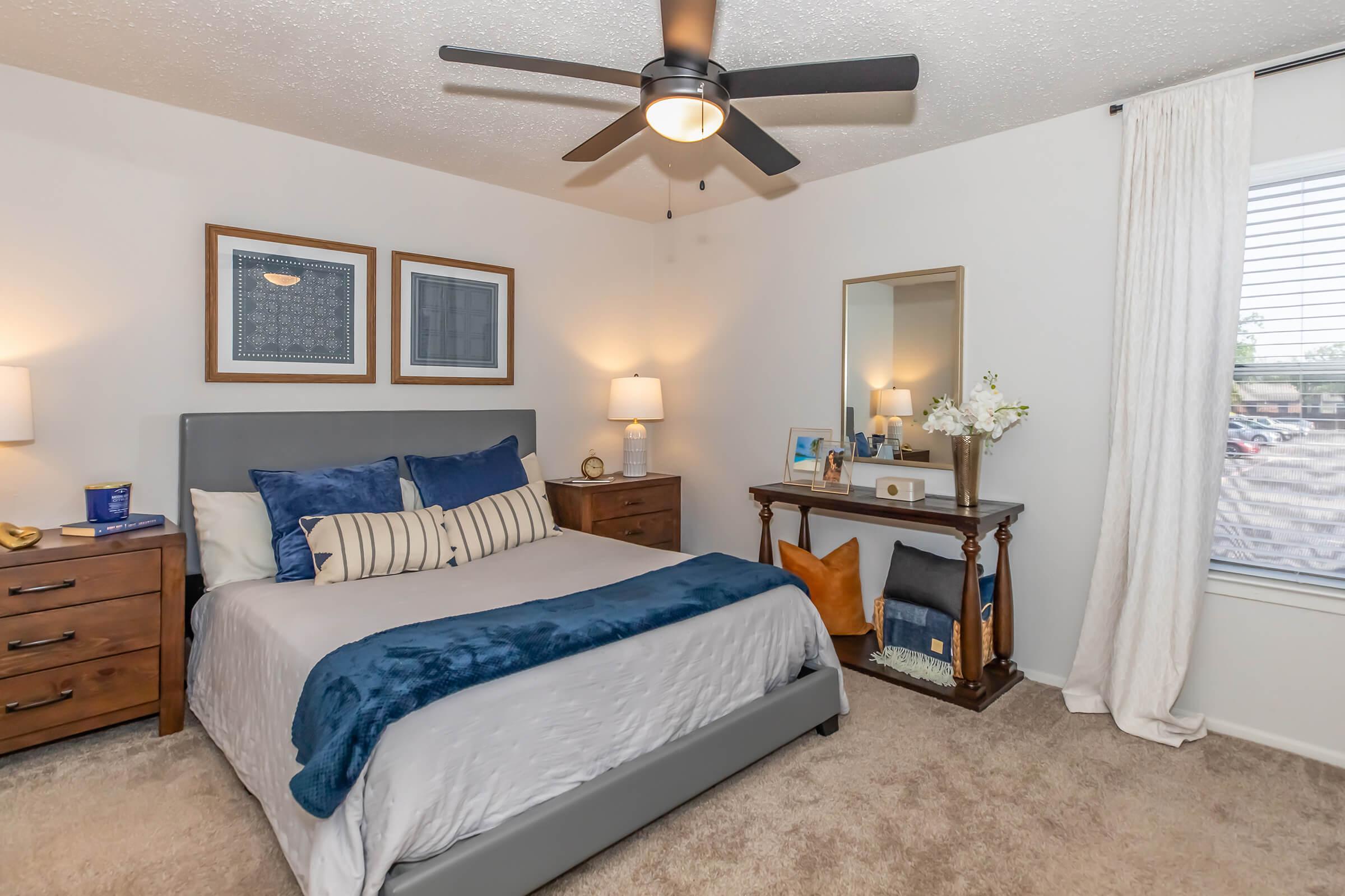 Cozy bedroom featuring a queen-sized bed with blue and white bedding, two bedside tables with lamps, a mirror, and a decorative table. Natural light filters through a window with sheer curtains, and a light-colored carpet adds warmth to the space.
