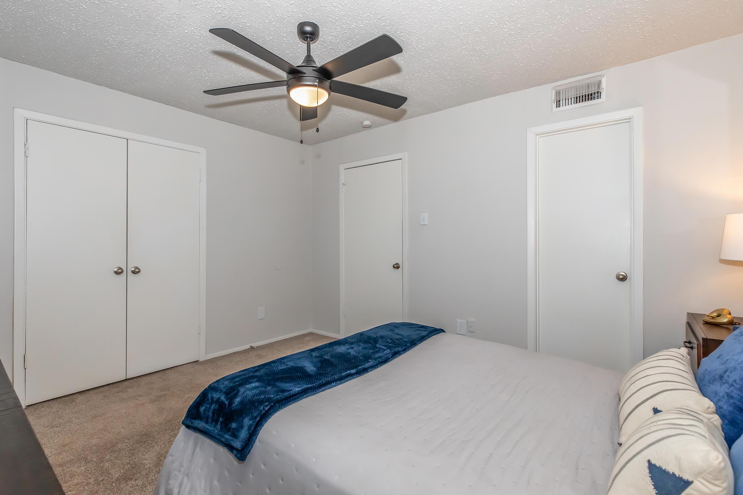 A cozy bedroom featuring a neatly made bed with a blue throw blanket, a nightstand with a lamp, and two closet doors. The walls are painted light gray, and there’s a ceiling fan above. The carpeted floor adds warmth to the space, creating a comfortable and inviting atmosphere.