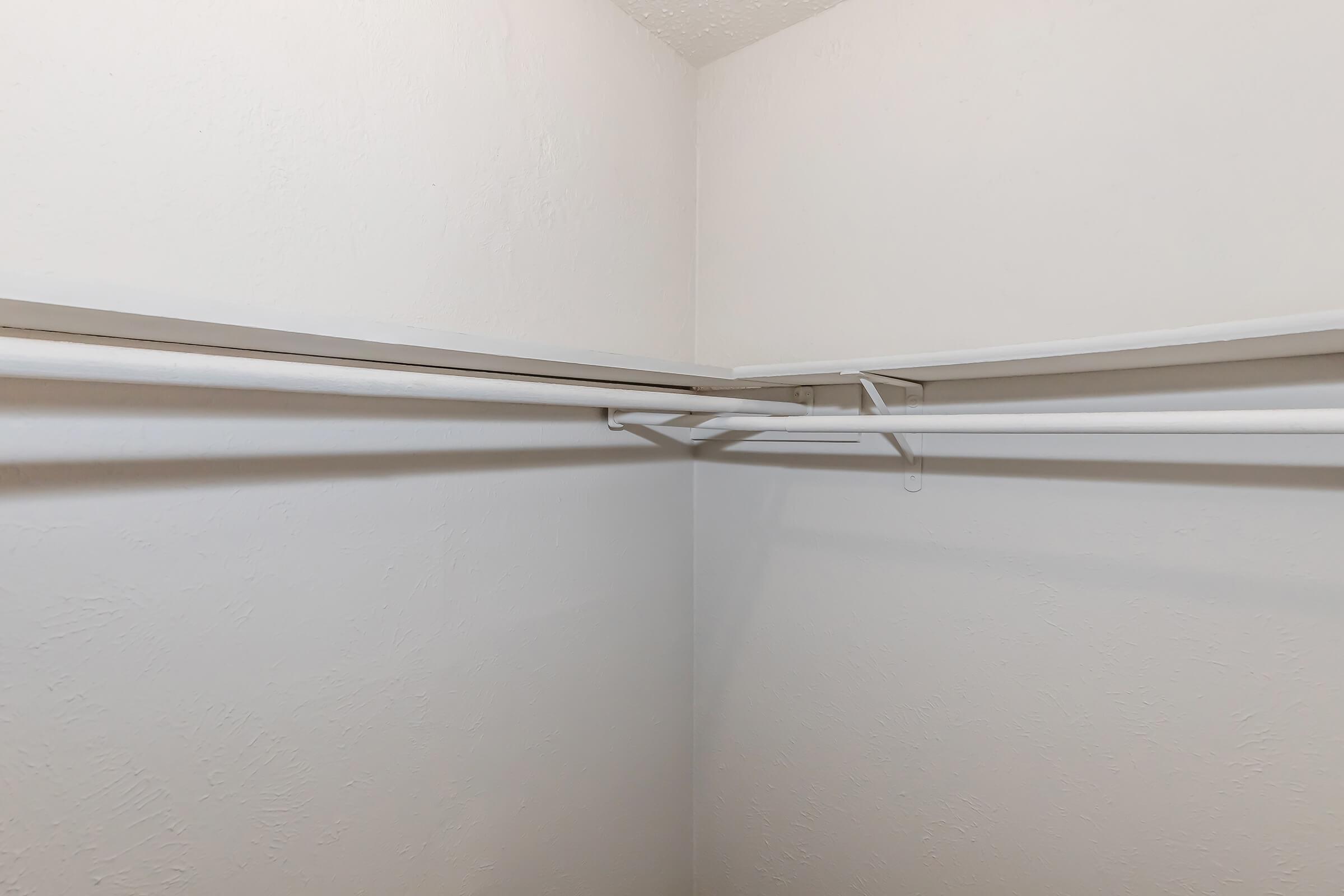 An empty closet corner featuring white walls and two horizontal closet rods. The rods are positioned on the upper part of the walls, showing no hanging items or shelves. The floor is visible, but the space appears tidy and organized.