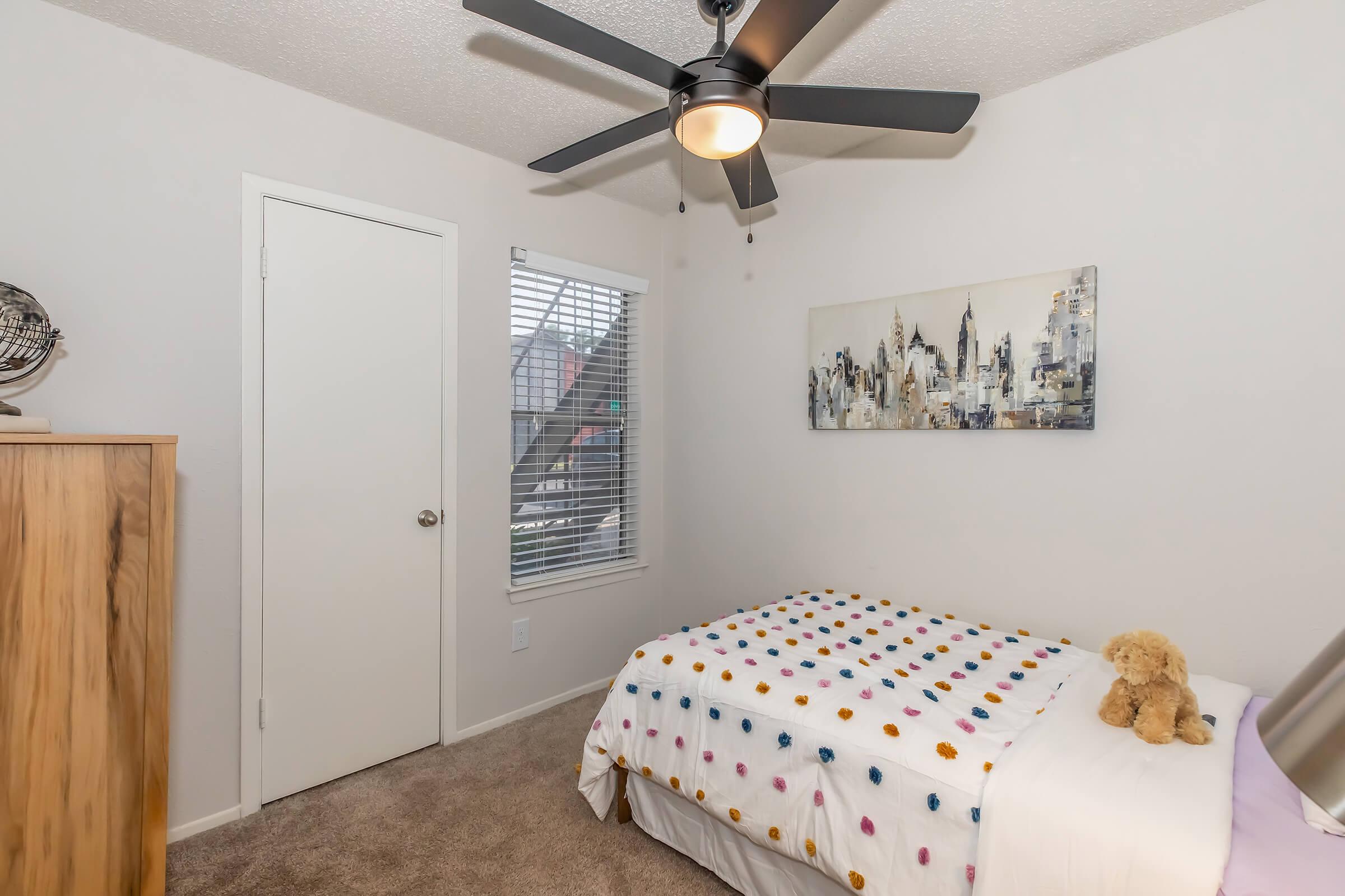 A cozy bedroom featuring a bed with a polka-dotted duvet cover, a stuffed teddy bear on the bed, a wooden wardrobe, and a wall art piece depicting a city skyline. The room has a ceiling fan and a window with blinds, allowing natural light to enter. A white door leads to another area.