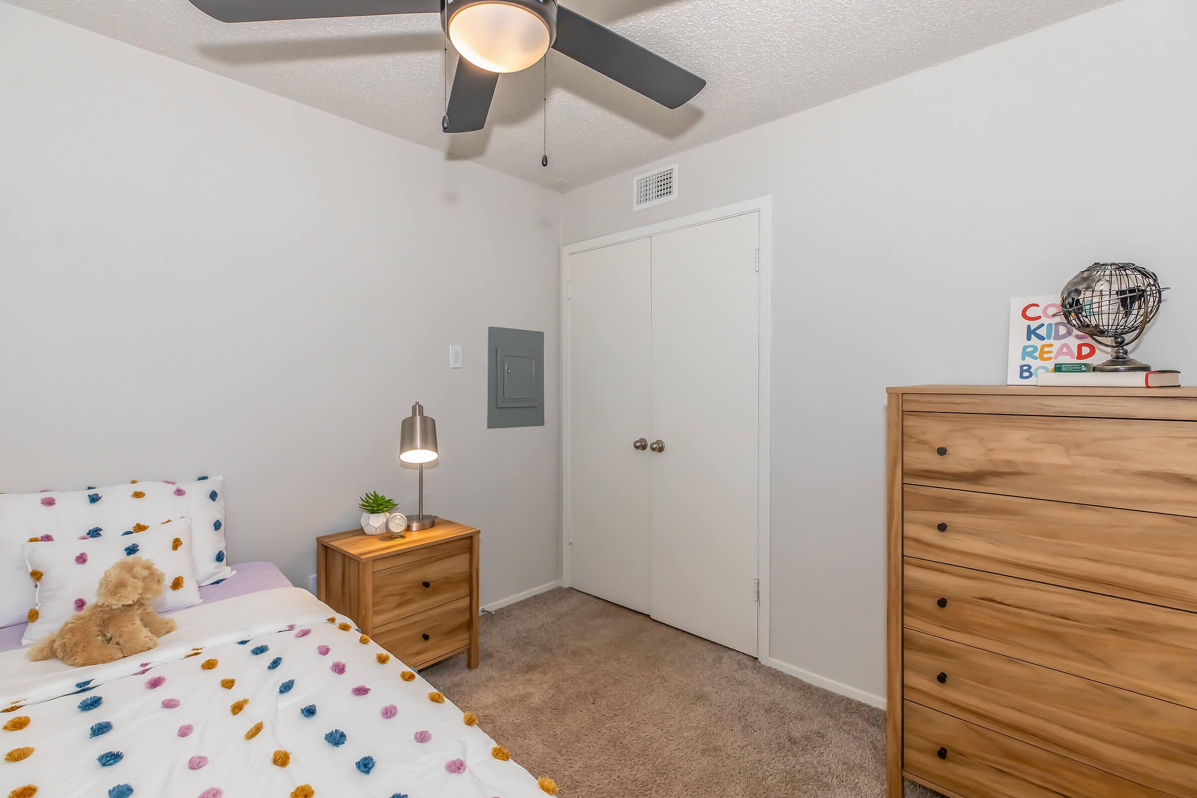 A cozy bedroom featuring a bed with a colorful polka-dot duvet cover, a wooden nightstand with a lamp, and a cute teddy bear. There is a dresser against the wall, and a ceiling fan above. The room has neutral-colored walls and a closet door in the background. A decorative book and globe are on the dresser.