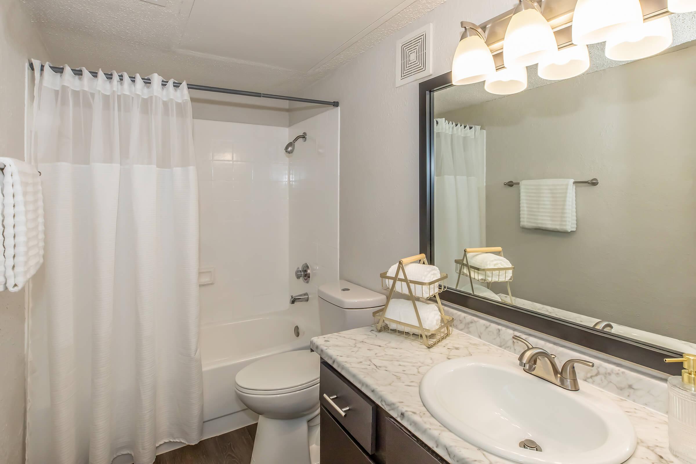 A compact bathroom featuring a white shower curtain, a bathtub with a showerhead, a modern toilet, and a sink with a marble countertop. Above the sink, there is a large mirror with three light fixtures. The bathroom has neutral-colored walls and a towel rack with a white towel.