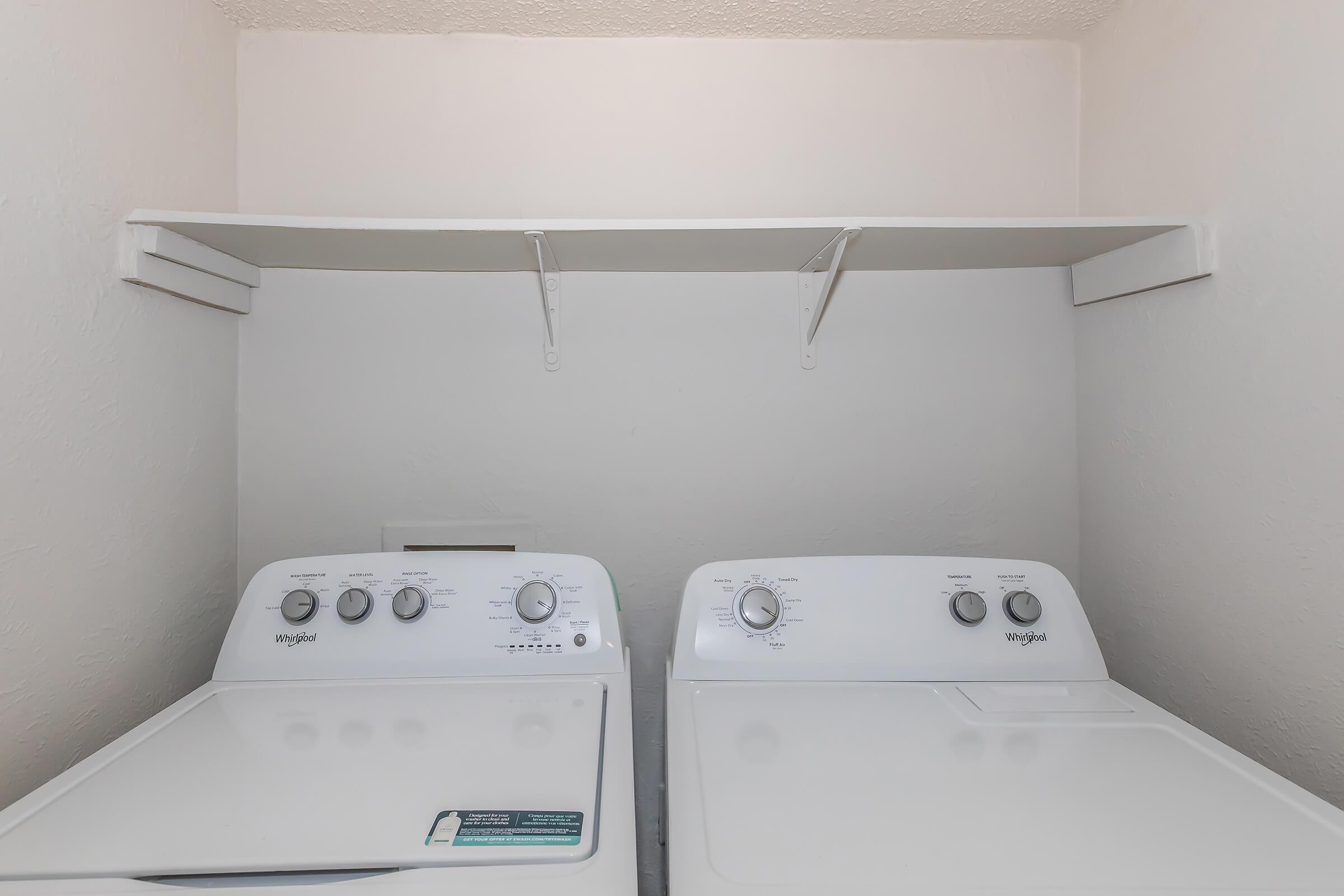 A laundry area featuring two white washing machines side by side. Above them, there is a simple white shelf mounted on the wall for additional storage. The space has a neutral, light-colored wall.