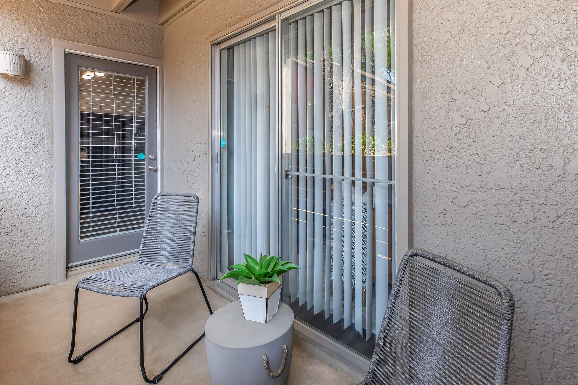 A cozy balcony featuring two modern chairs and a round side table with a small potted plant. The sliding glass doors have vertical blinds, allowing light to enter while maintaining privacy. The textured walls add a touch of elegance to the space.