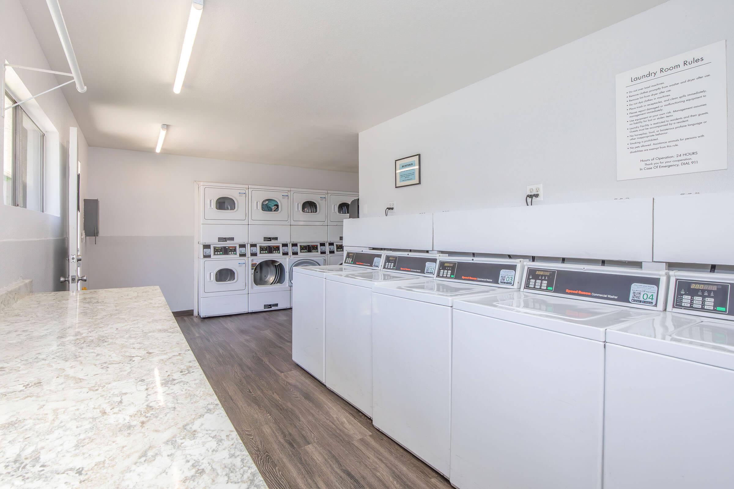 A clean and bright laundry room featuring several white washing machines and dryers against a light-colored wall. There’s a marble countertop for folding clothes and a sign with laundry room rules on the wall. Large windows allow natural light to fill the space, creating a welcoming environment.
