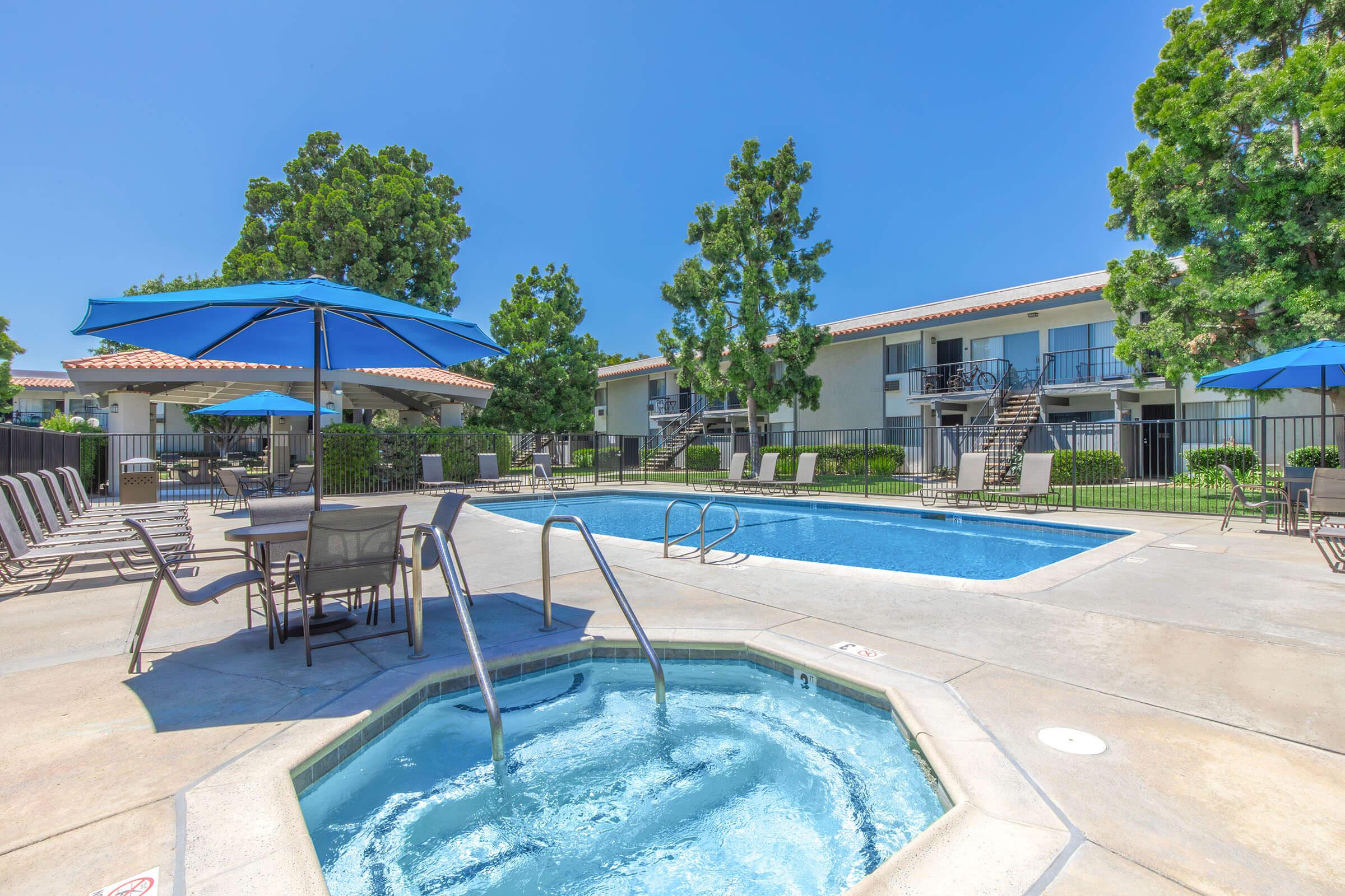 A sunny outdoor pool area featuring a clear blue swimming pool and a hot tub. Surrounding the pool are lounge chairs and umbrellas for shade, with lush trees in the background and two-story apartment buildings visible in the distance. The scene conveys a relaxing atmosphere perfect for leisure.