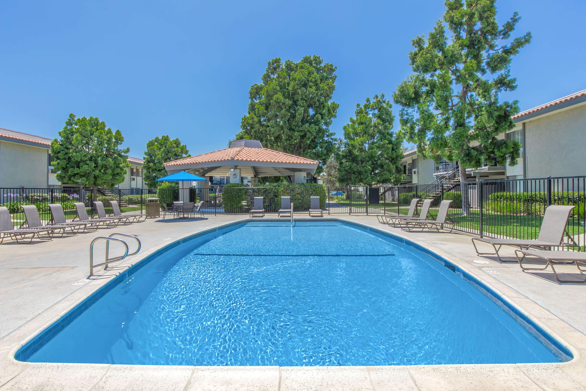 A clear blue swimming pool surrounded by lounge chairs, with a gazebo and shaded seating area nearby. Lush green trees provide a backdrop, and the pool area is fenced. The scene is bright and sunny, indicating a warm day, with apartment buildings visible in the background.