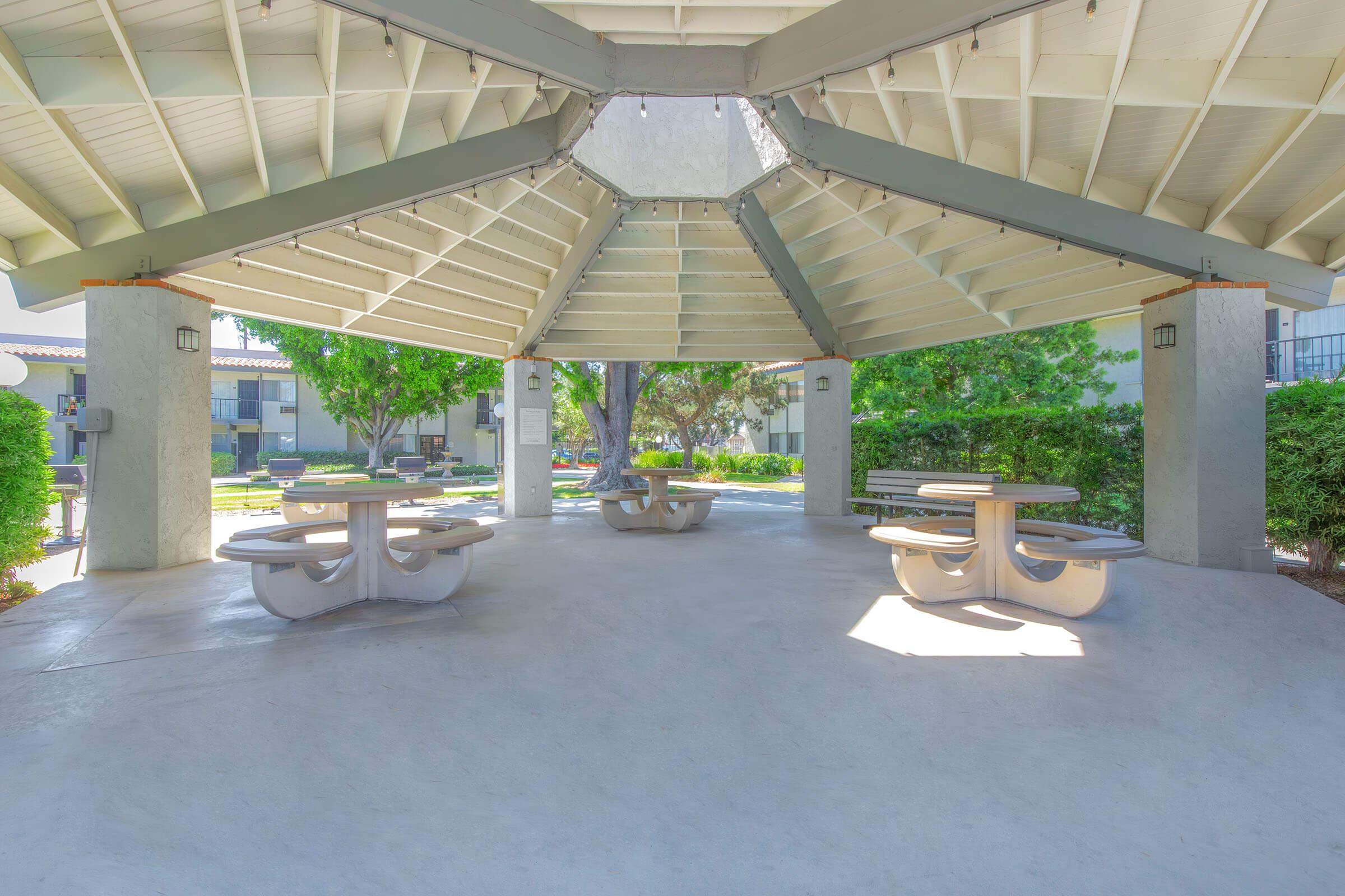 A covered pavilion with a large, circular seating area featuring multiple picnic tables. The space is surrounded by green trees and well-maintained landscaping, creating a serene outdoor atmosphere. The roof is supported by gray beams, and the floor is smooth concrete. Ideal for gatherings or relaxing.