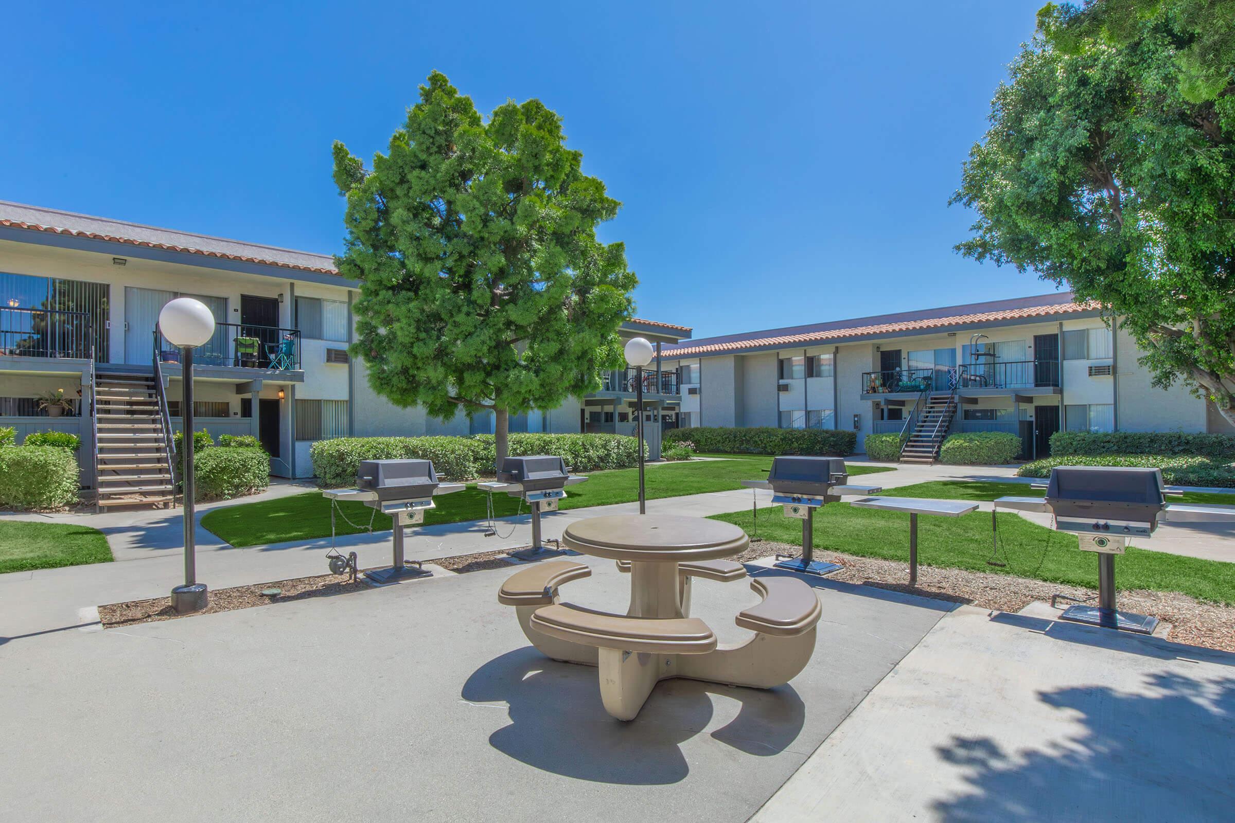 A bright outdoor space featuring a round picnic table surrounded by grill stations and well-maintained landscaping. Apartment buildings with balconies are visible in the background under a clear blue sky, creating a welcoming environment for gatherings.