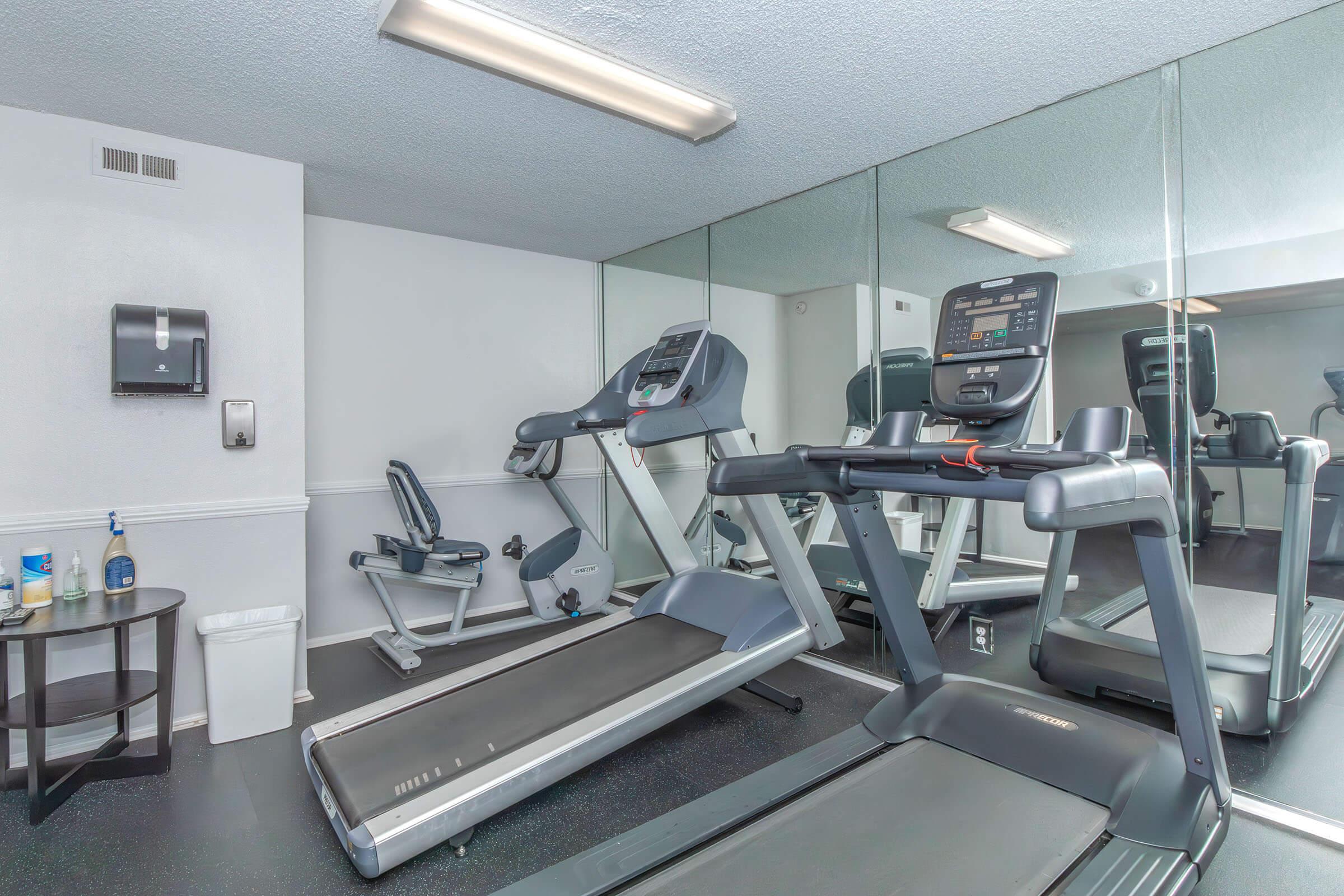 A well-lit gym area featuring two treadmills, a stationary bike, and a small table with fitness supplies. The room has mirrored walls and a clean, modern design, suggesting a space for physical activity and exercise.