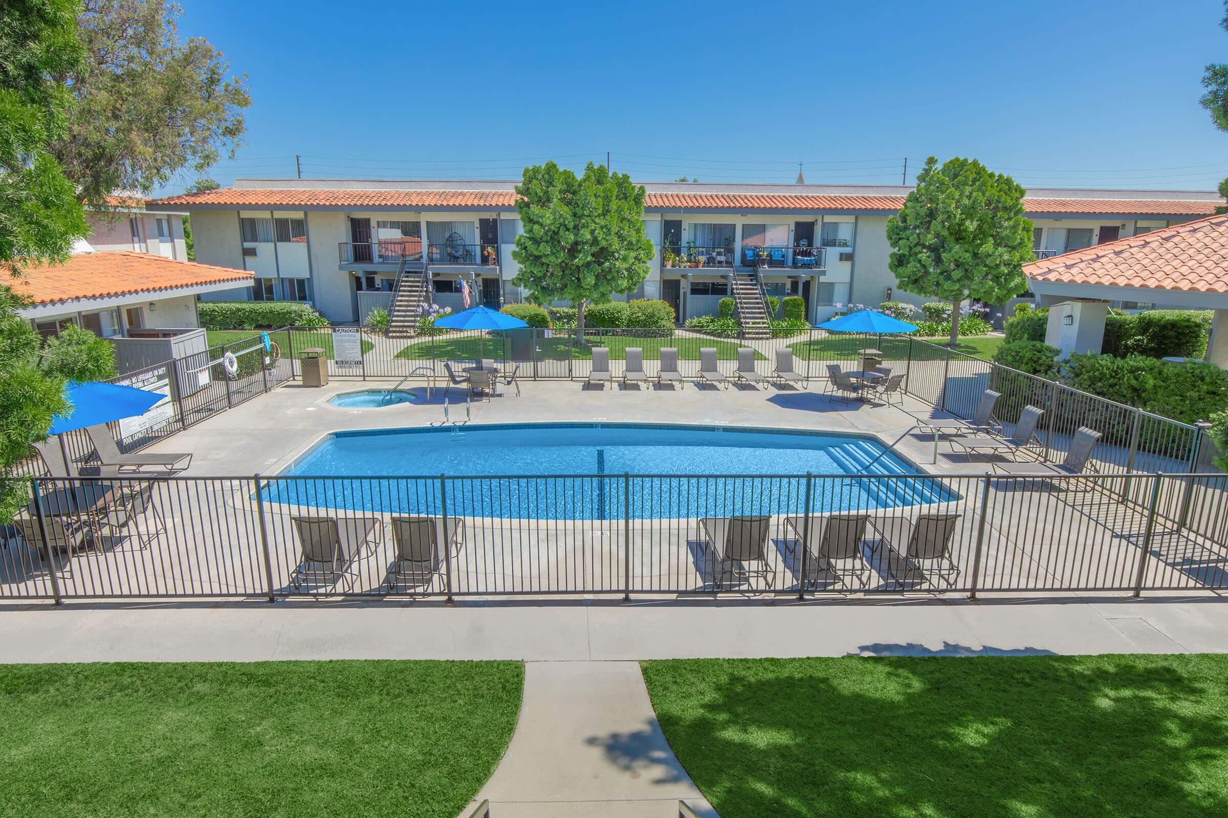 A swimming pool surrounded by lounge chairs and blue umbrellas, with landscaped greenery and two-story buildings in the background. The sky is clear and bright, indicating a sunny day.