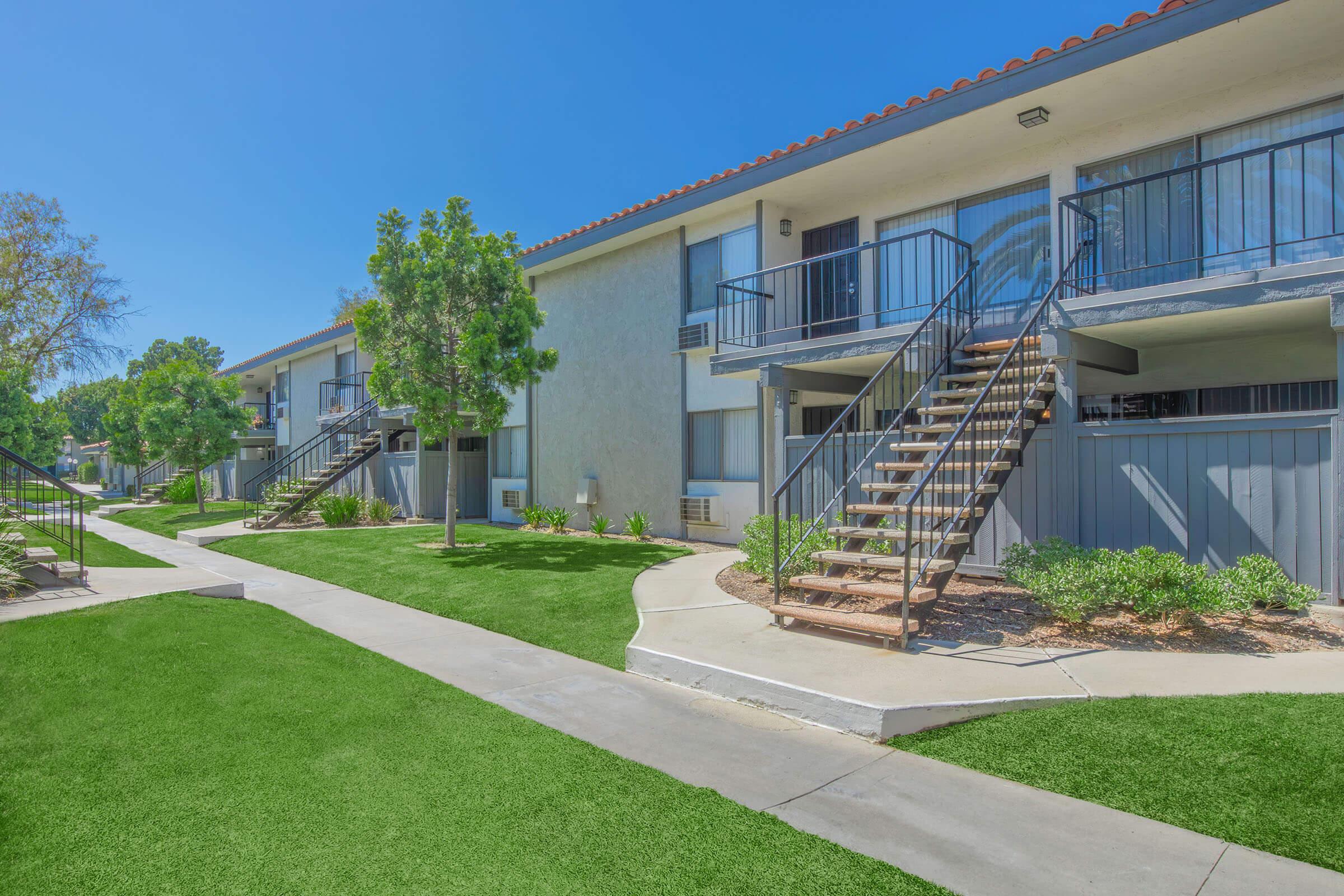 A view of a multi-unit residential building featuring two staircases, greenery, and well-maintained pathways. The buildings have a light exterior with balconies and are surrounded by manicured lawns under a bright blue sky.