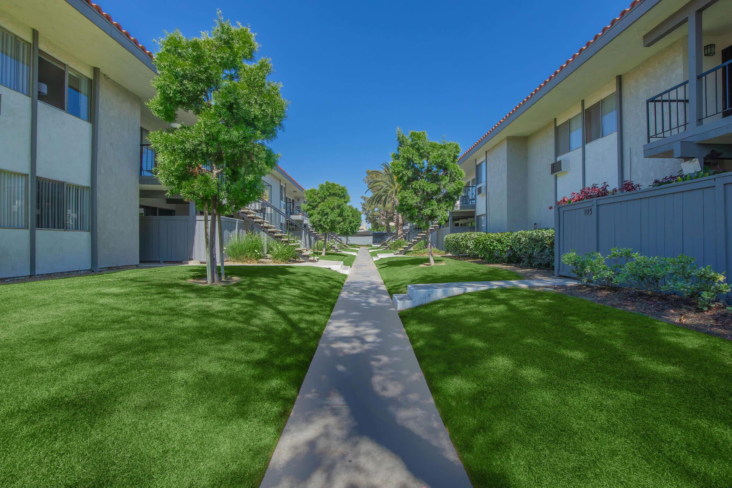 A well-maintained apartment complex with two-story buildings on either side of a paved walkway. Lush green lawns and neatly trimmed shrubs and trees enhance the landscape under a clear blue sky, creating a welcoming and serene outdoor environment.