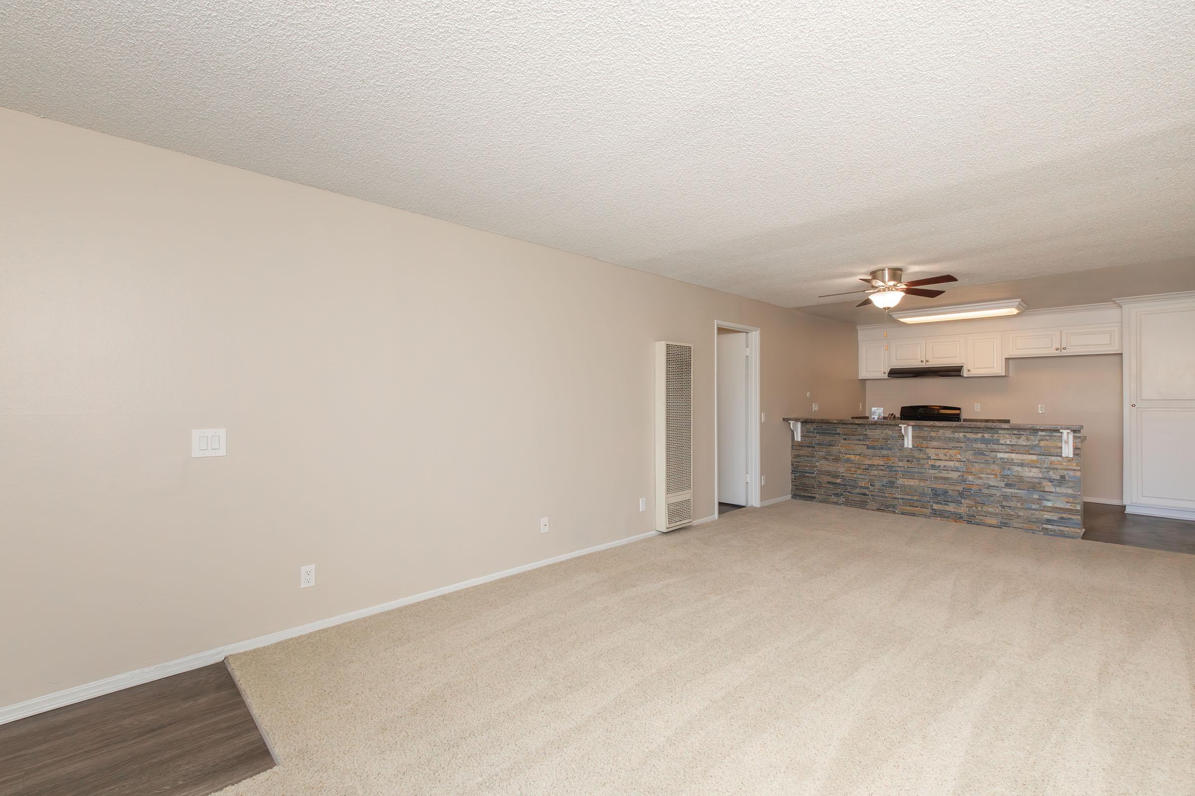 Spacious living area with light beige walls and a textured ceiling. The floor is covered with a soft beige carpet. In the background, there's a kitchen area featuring a stone bar counter. A ceiling fan is mounted above the lounge space, and a small white pantry is visible on the left side.