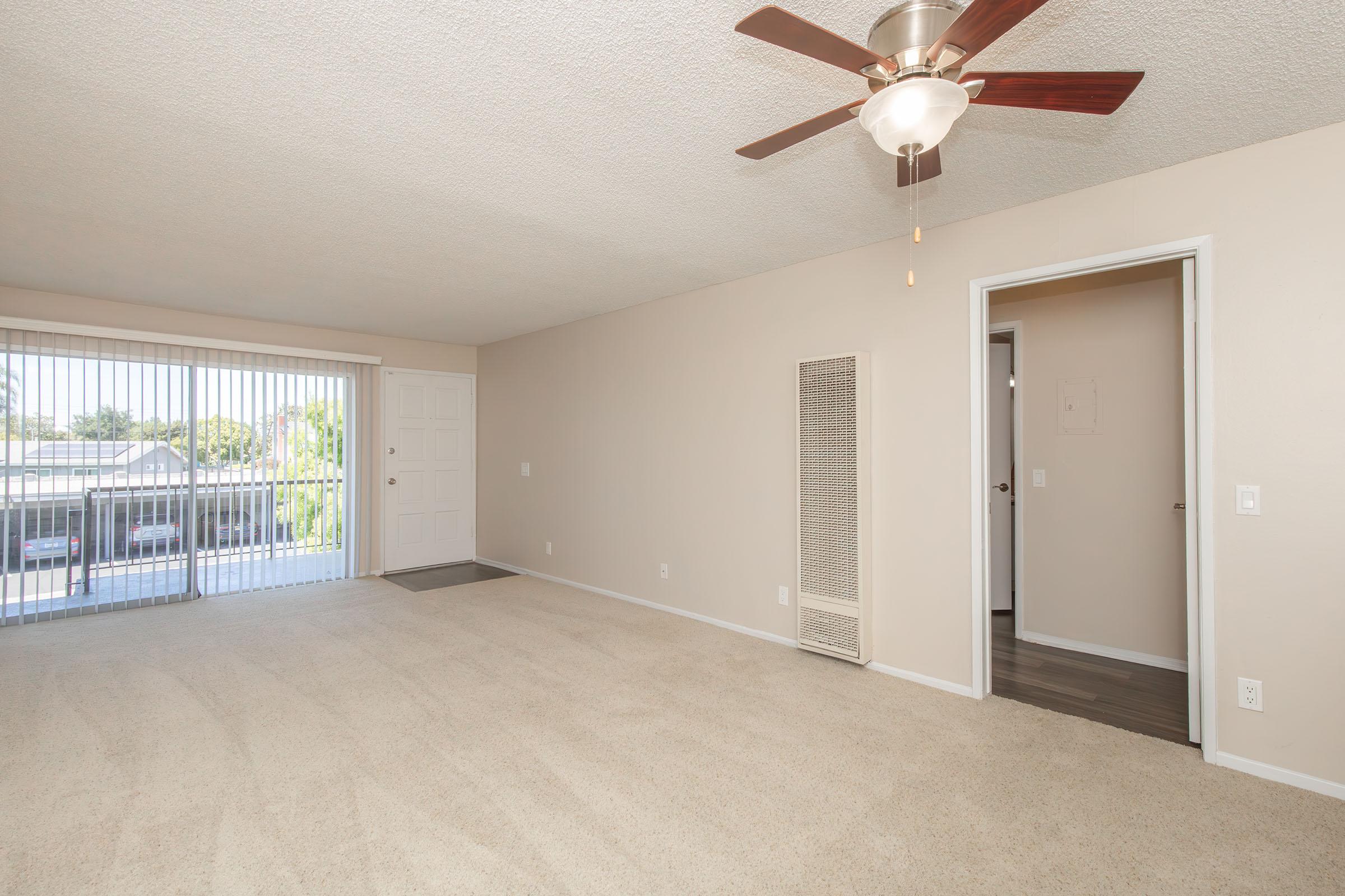 A spacious, light-filled room with beige carpet and a ceiling fan. One wall features a sliding glass door leading to a patio, while a nearby door leads to another room. The walls are painted a soft beige, creating a warm ambiance. A vent is visible on the right side of the room.