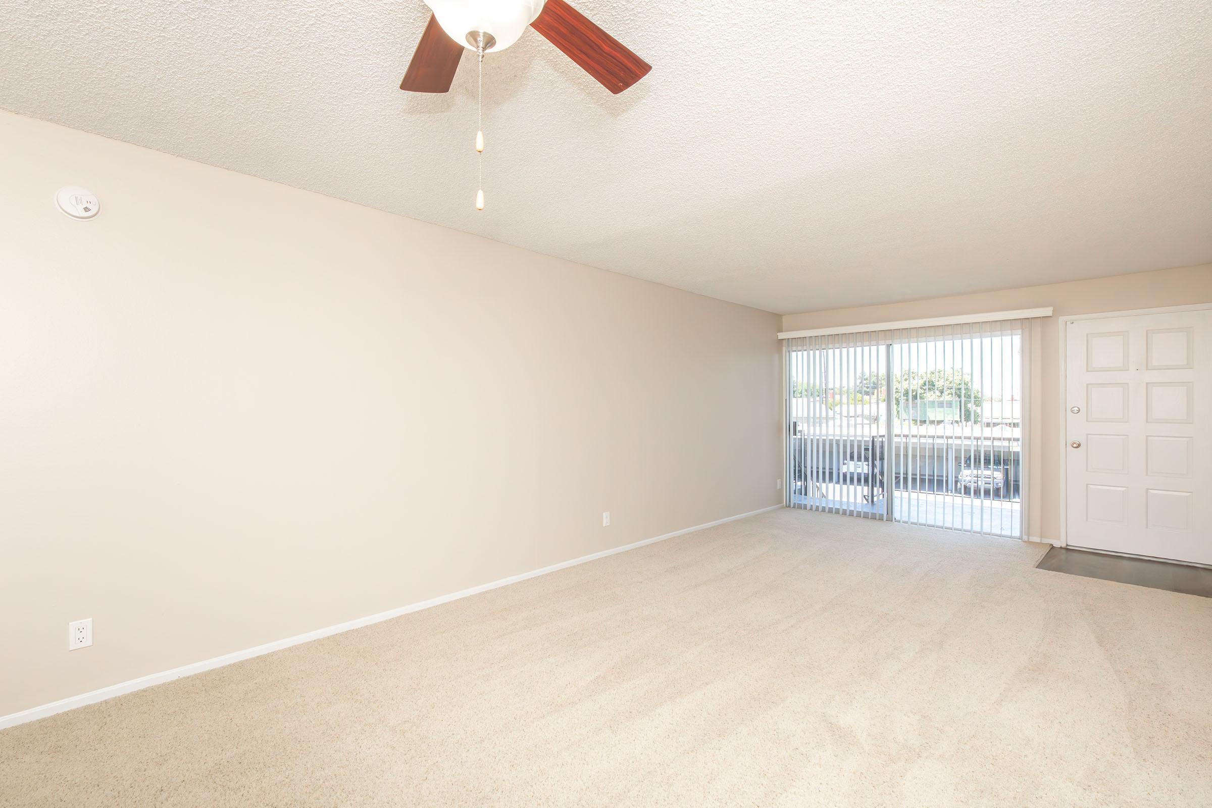 Spacious living room featuring beige walls and carpet, with a ceiling fan. A sliding glass door leads to an outdoor area, allowing natural light to enter. The entrance includes a white door and a doormat, creating a welcoming atmosphere.