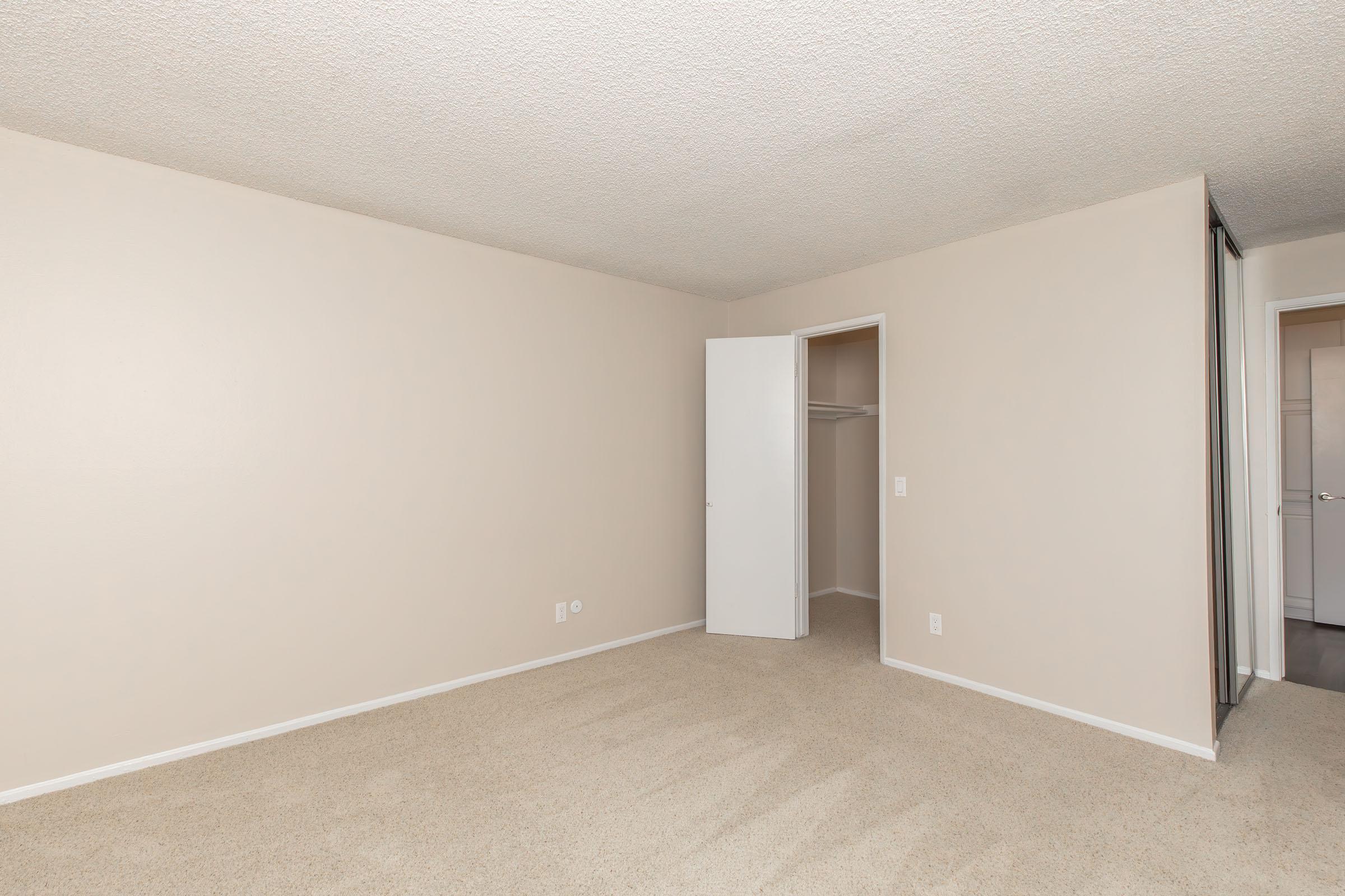 Empty room with beige walls and carpeted floor. A white closet door is slightly open on the right, revealing a space with no contents. Natural light is coming from an unseen window, giving a neutral and spacious feel to the area. No furniture or decorations are present.