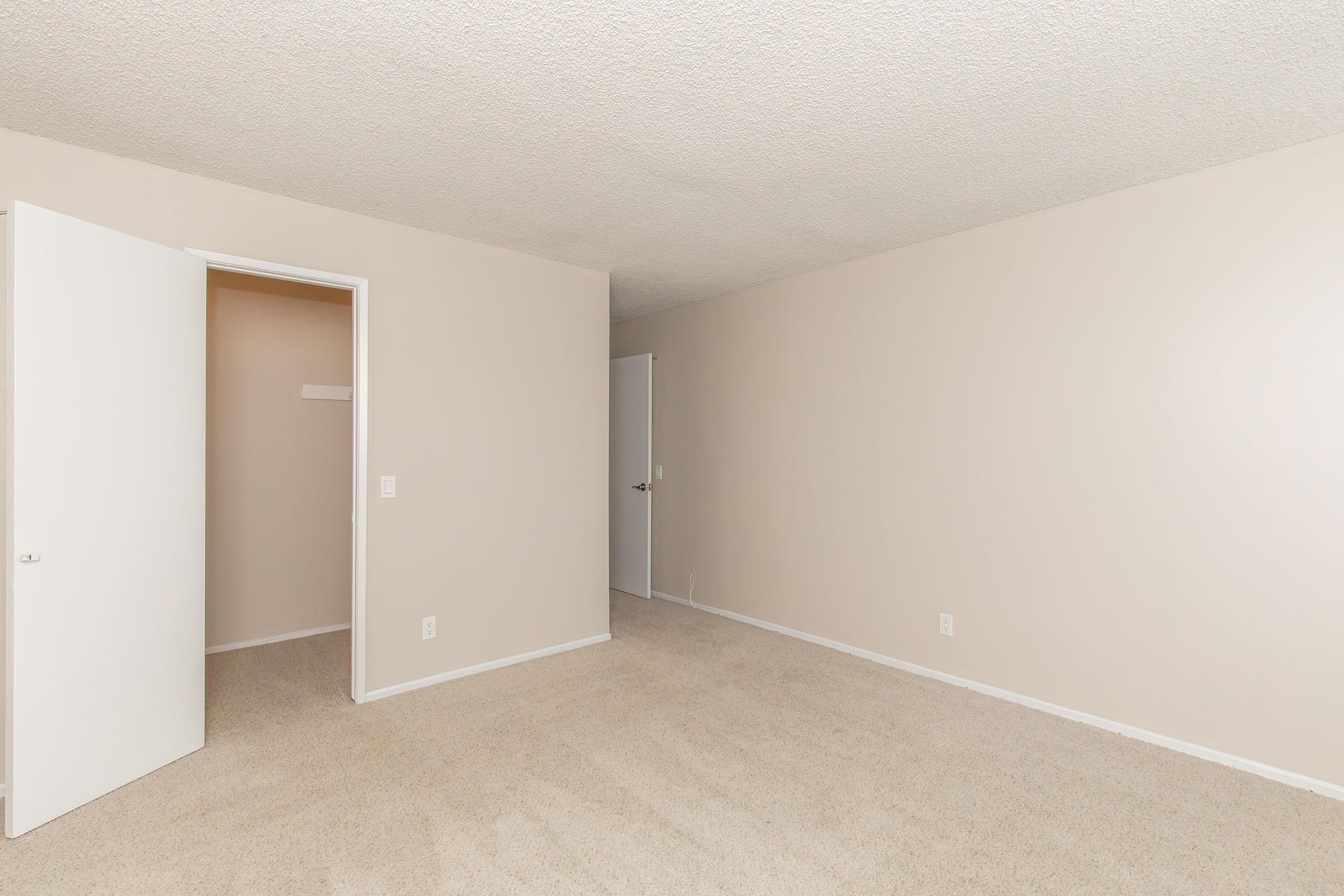A well-lit, empty room with beige walls and light carpet. There are two white doors: one open leading to a closet and the other closed, indicating another room. The overall ambiance is clean and spacious, suitable for furnishing or decoration.