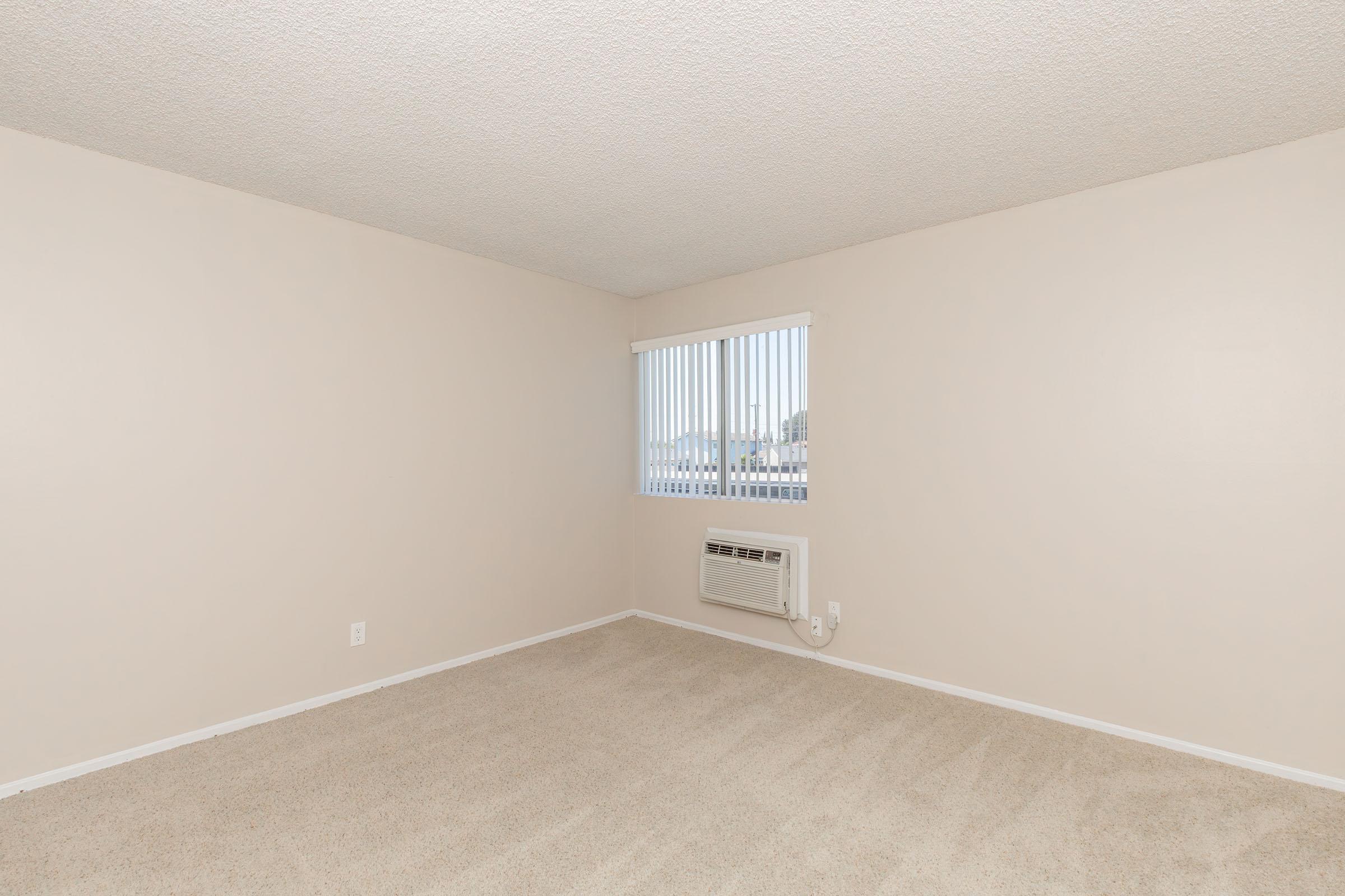 Empty room with beige walls and carpet, featuring a window with vertical blinds and an air conditioning unit on the wall. The room has no furniture and is well-lit, giving it a spacious feel.