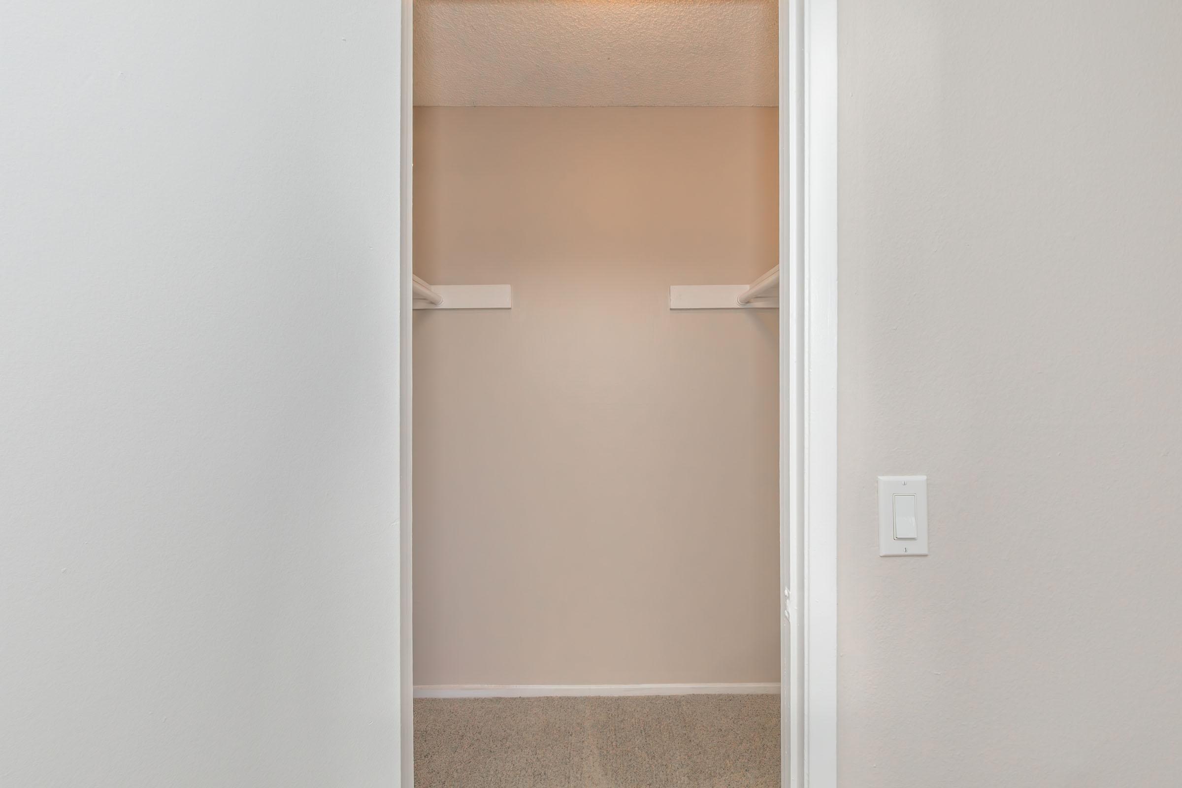 A clean, empty closet with light beige walls and a carpeted floor. Two shelves are visible on either side, and the doorway is framed with a white door trim. A light switch is located near the entrance. The overall atmosphere is bright and spacious.