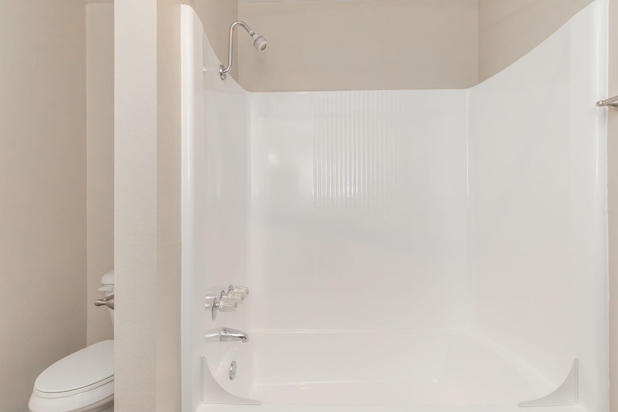 A clean, modern bathroom featuring a white, spacious shower with smooth walls. A shower head is mounted on the wall, and a white toilet is visible to the left, against a light-colored wall. The overall design is simple and minimalistic.