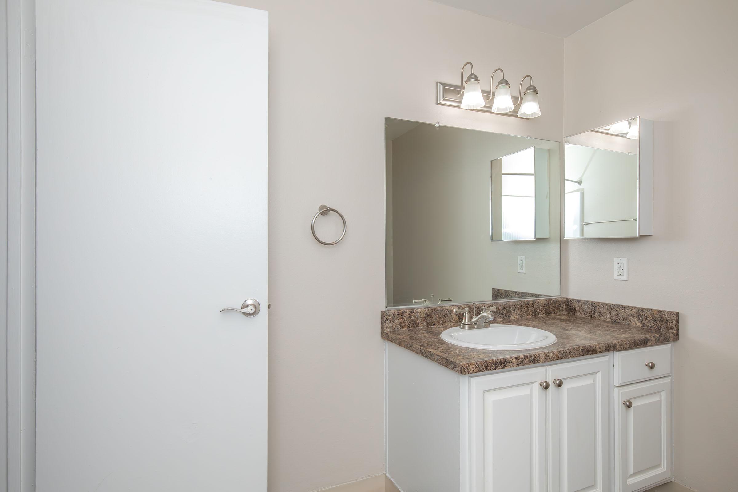A clean and simple bathroom featuring a white cabinet with a granite countertop, a round sink, a mirror, and wall-mounted lighting. The walls are painted in a light color, and there is a towel bar next to the sink. A white door leads to another space, creating a bright and spacious feel.