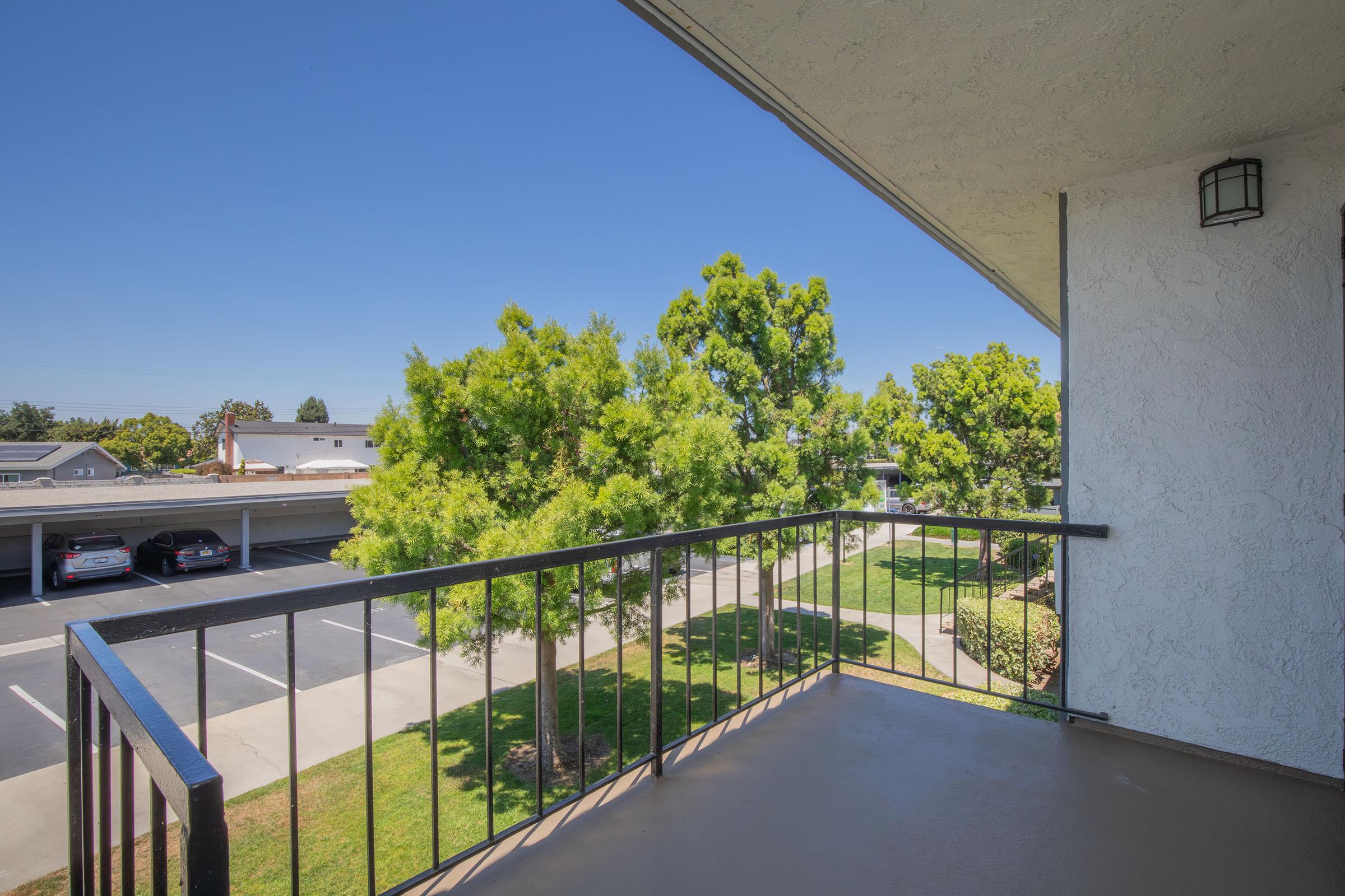 A view from a balcony overlooking a well-maintained landscape with trees and a parking area. The sky is clear and blue, and the surrounding buildings are partially visible. The scene conveys a sense of tranquility and openness.