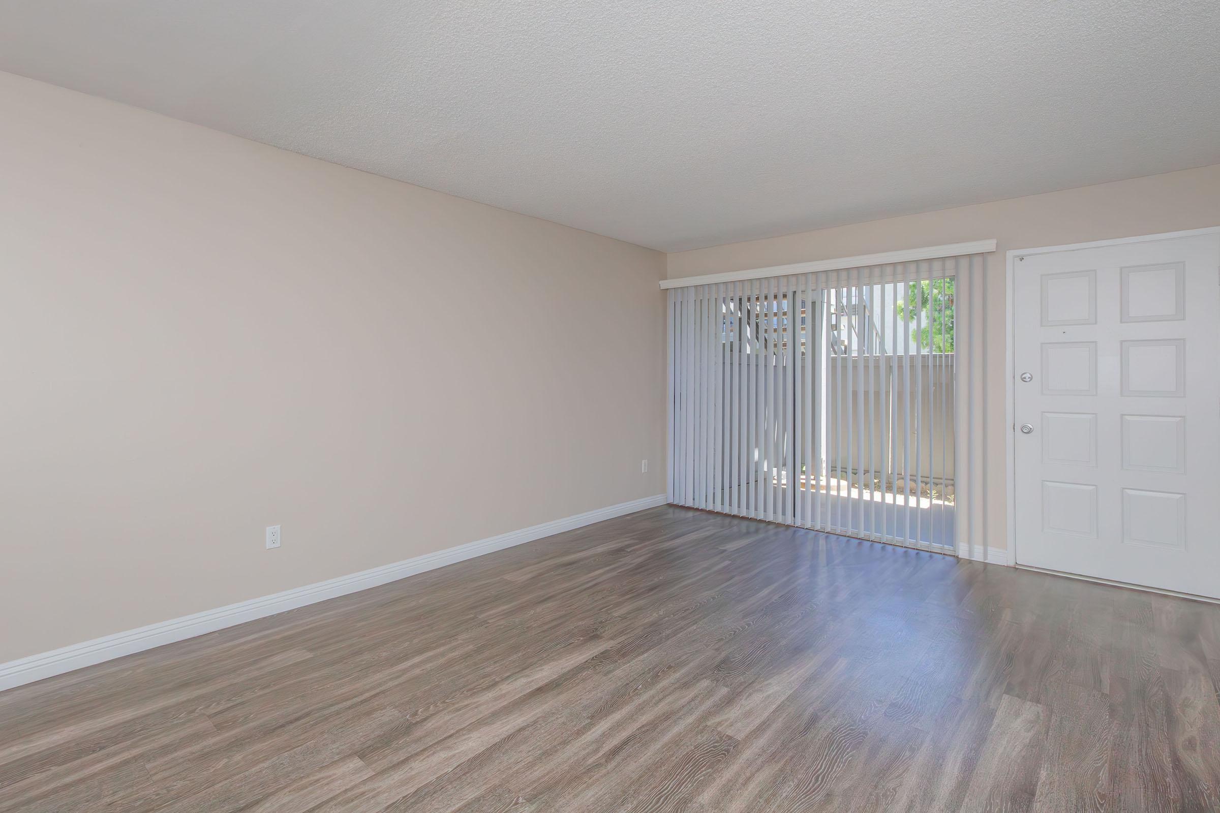 A light, empty room with a beige wall and wooden flooring. There are vertical blinds on a large window facing an outdoor space. A white door is visible on one side, contributing to the minimalist and airy atmosphere of the room.